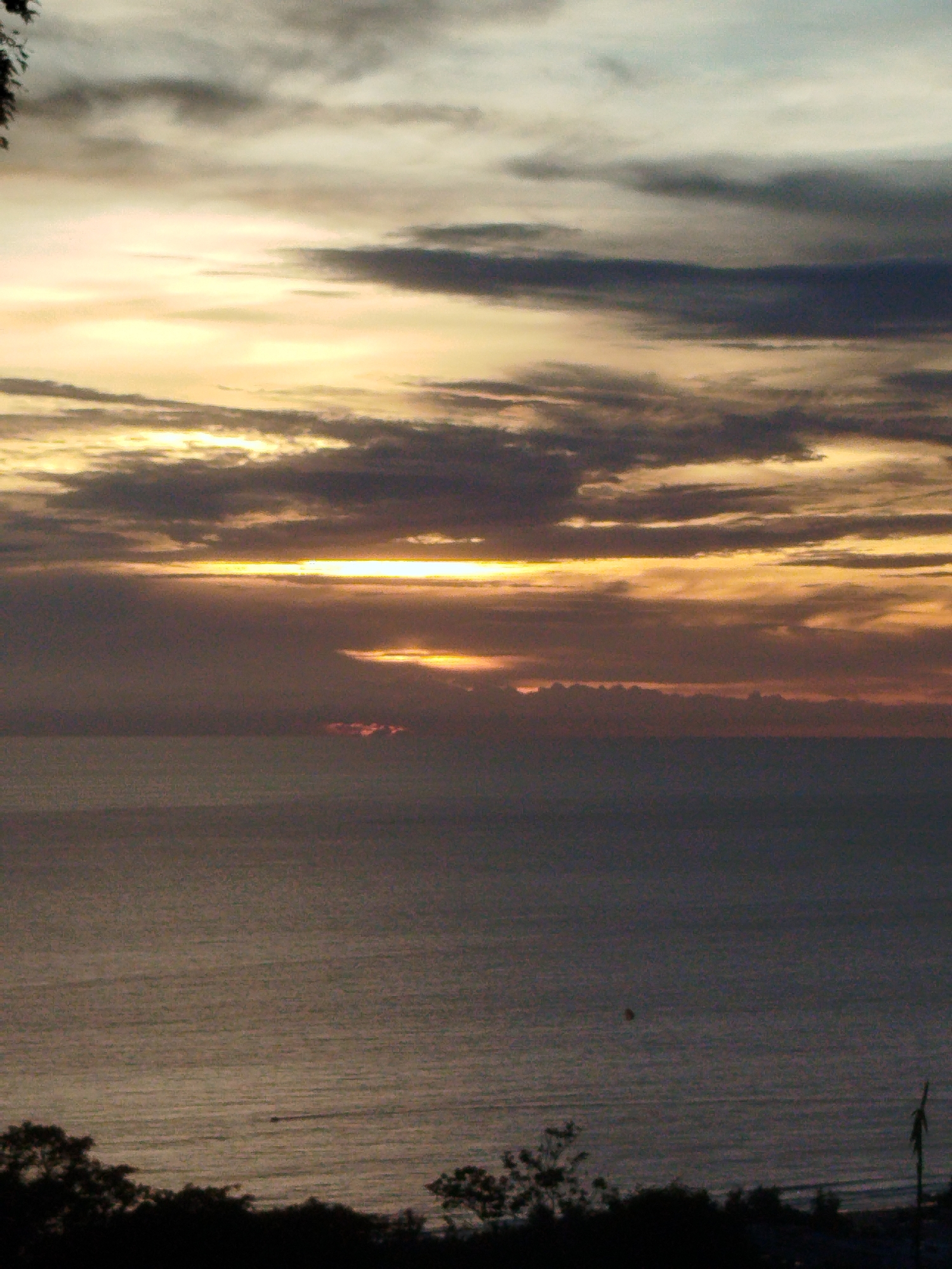 The image depicts a serene seascape at dusk, likely in Karon, Thailand. The sky is filled with dramatic clouds, painted in hues of orange, yellow, and dark gray as the sun sets or rises. A prominent horizontal band of bright, warm light streaks across the middle of the sky, suggesting the sun is just below the horizon. Below the sky, the vast ocean stretches out, its surface reflecting the subdued light. The water appears calm with subtle ripples. In the foreground, the dark silhouettes of lush trees and foliage frame the bottom of the scene, creating a stark contrast with the illuminated sky and water. The overall atmosphere is one of tranquility and natural beauty. The time of day is clearly sunset or sunrise, given the quality of the light and the cloud formations. The presence of a shoreline and abundant vegetation points to a coastal location.