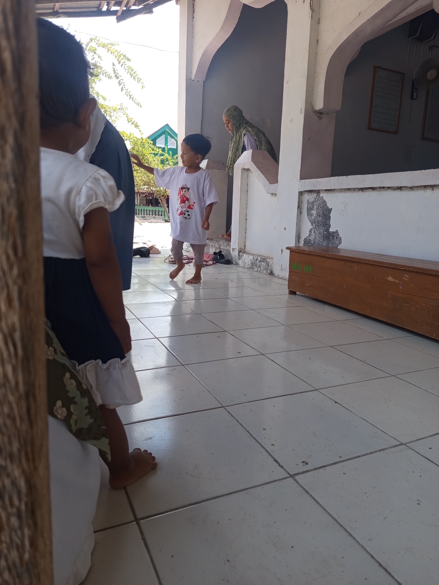 In Lumutan, Indonesia, a young boy in a white t-shirt with a graphic of a soccer player and a soccer ball is standing on a tiled floor, extending his right hand towards the left. He is barefoot and appears to be engaged with another child whose back is to the camera. This child is wearing a white top and a dark skirt with ruffled trim. In the background, a woman in a green headscarf is partially visible looking down. The setting appears to be an outdoor covered area with a pale pink arched structure and a white wall. To the right, a long, brown wooden bench with green lettering, possibly "MANF", is present. The floor is tiled in a light color with visible grout lines. Sunlight illuminates the scene, creating shadows on the floor and walls.