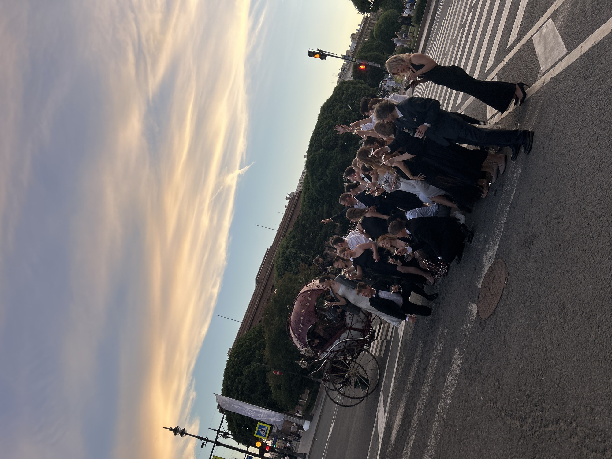 In an outdoor urban setting, a group of individuals dressed in formal attire, including tuxedos and gowns, are posing for a photograph. They are gathered around a horse-drawn carriage, with many holding champagne glasses. The scene takes place on a crosswalk, with a traffic light visible in the background. The sky displays a dramatic sunset with clouds painted in hues of orange and yellow. Trees and buildings form the backdrop.