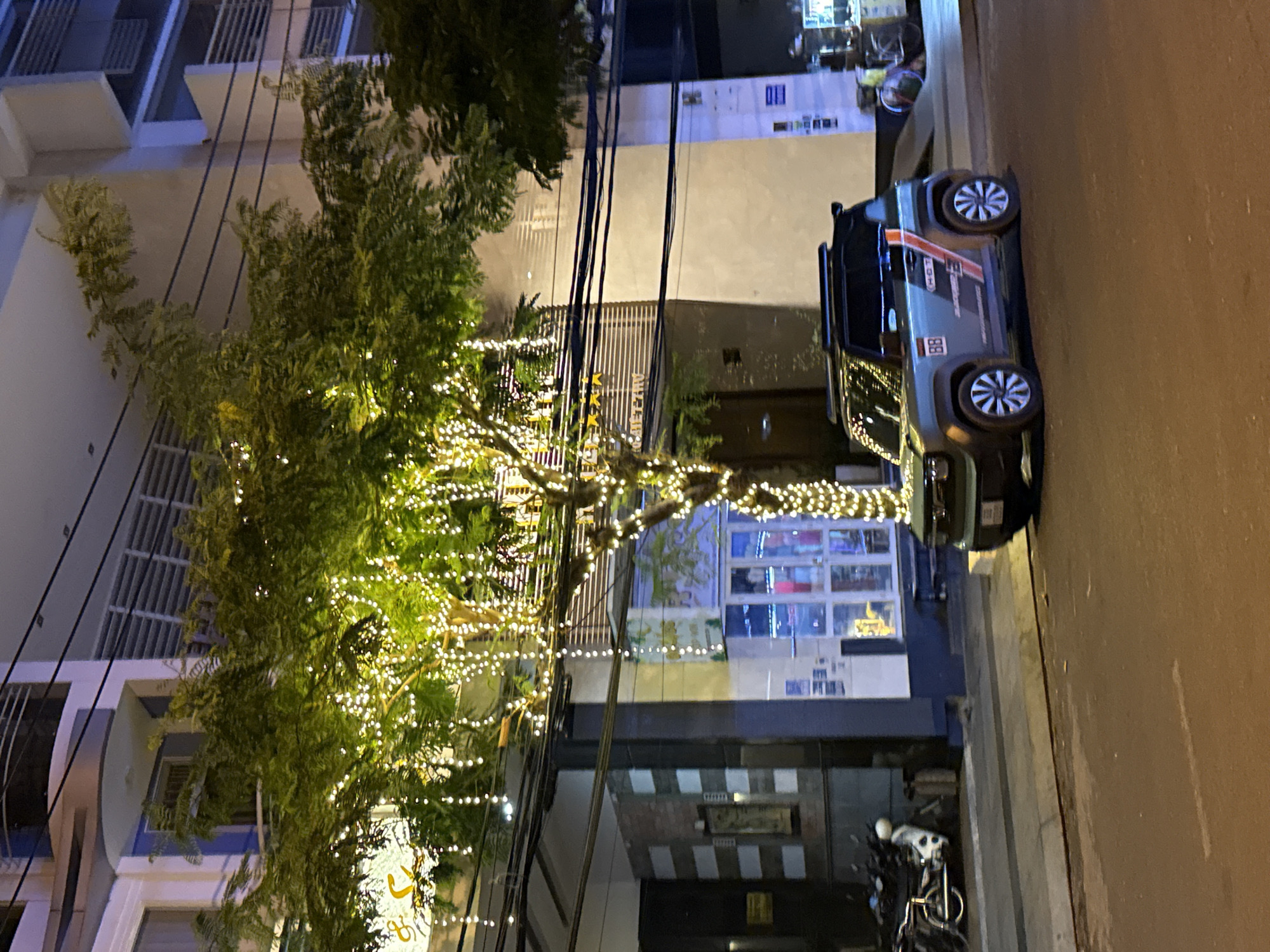 The image depicts a street scene in Nha Trang Ward, Vietnam, at night. A tree heavily decorated with white lights stands on the street, partially obscuring the view of buildings across the street. Electrical wires crisscross the scene above the tree. Parked on the street is a gray vehicle with a red and white stripe design. A motorcycle is parked nearby. The street is illuminated by streetlights and lights from the buildings. The buildings appear to be a mix of residential and commercial establishments.