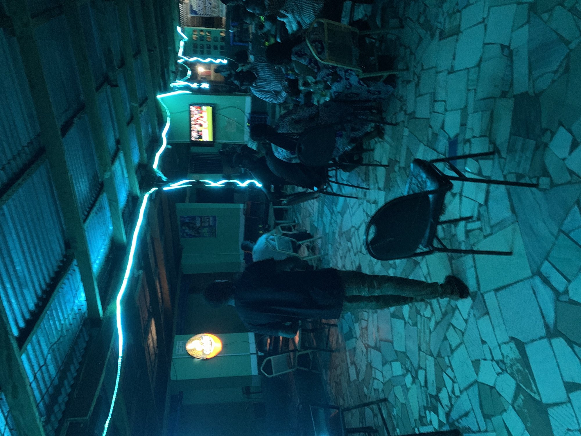 This is a nighttime image of what appears to be an outdoor or semi-outdoor bar or entertainment venue located in or near New Stadium, Nigeria. The image features a group of people seated in the background, some watching a television screen. A man is walking or standing in the foreground. The area is lit with blue neon lights lining the corrugated metal roof, and there is also a circular light fixture on one of the walls. The floor appears to be tiled with irregular shapes. The overall atmosphere is casual and dimly lit.