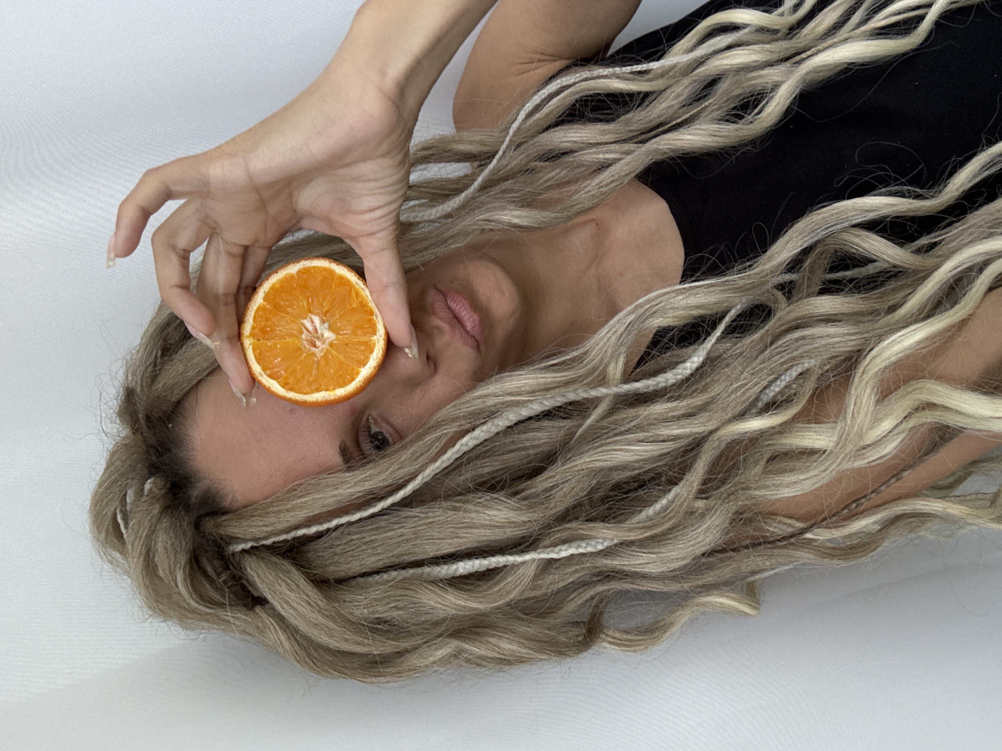 A person with light brown, wavy hair intermixed with thin white braids is lying on a white surface. They are wearing a black top. Their right hand holds a halved orange slice in front of their right eye. The location in the city of Rusanovo, Russia, cannot be determined from this image.