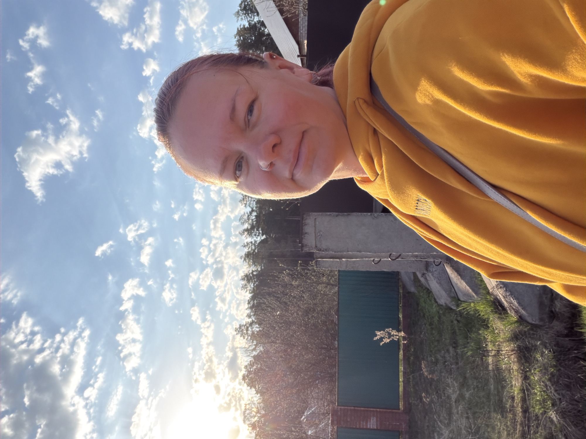 A woman wearing a mustard-colored hooded sweatshirt is in the foreground. The background includes a blue sky with scattered clouds, trees, a green fence, a concrete structure, and grass. The location is possibly in the city of Chaikovsky, Russia. The sun is near the horizon.