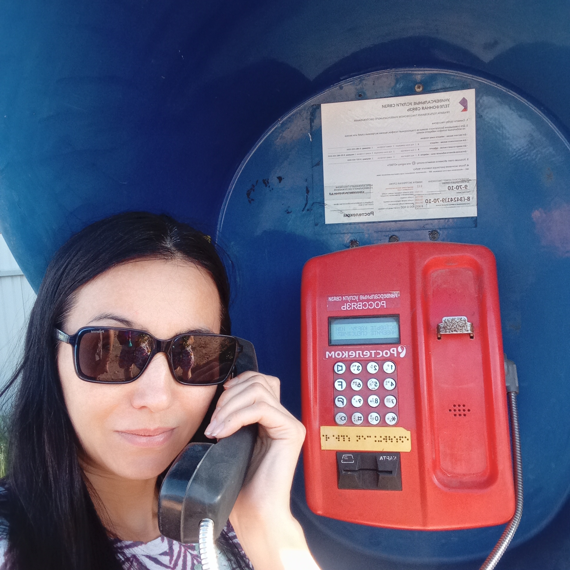 A woman with dark hair and sunglasses is using a red payphone in a blue phone booth. The phone booth contains a notice with Russian text, and the payphone keypad includes Russian characters and braille. The payphone displays Cyrillic characters. The booth is in the city of Буренка, Russia.
