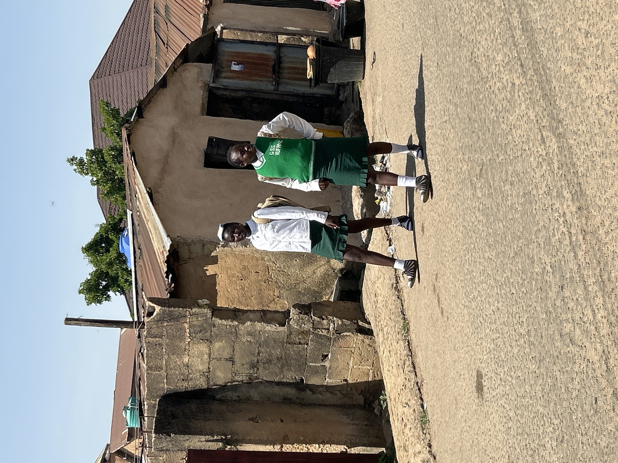 Two dark-skinned girls, presumed to be students, are standing on a concrete road. They are wearing white, long-sleeved shirts, green vests with "GSS HUFFING" printed on them, and dark green skirts. Both have on white socks and black and white sneakers. They are positioned in front of a dilapidated structure with a tin roof. The structure appears to be made of a combination of brick and mud. The background shows other buildings with tin roofs. The scene is most likely located in a low-income area in Jos, Nigeria.