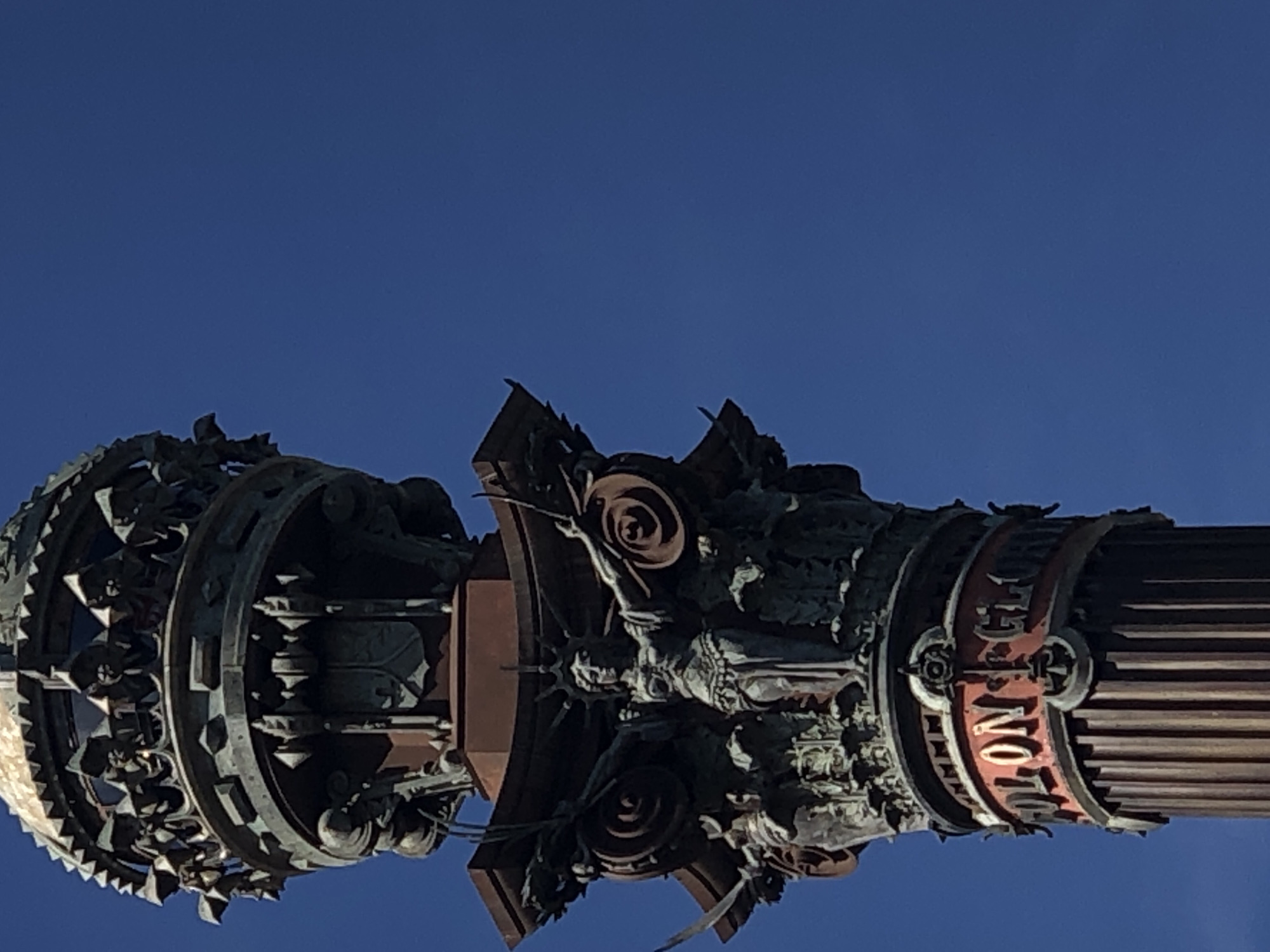 The image shows a close-up, low-angle view of the top section of a large, ornate monument, likely a column, set against a clear, vibrant blue sky. The monument appears to be made of bronze or a similar dark metal, with some reddish-brown elements.
The uppermost part of the visible monument features a crown-like structure with intricate detailing, possibly incorporating small figures or decorative motifs. Below this, a prominent band of intricate carvings and decorative elements is visible, including what appears to be a shield with a crest.
Further down, a central female figure is depicted with outstretched arms, wearing a crown and a flowing garment. She is surrounded by further sculptural work, including what look like scroll-like elements and possibly allegorical figures.
Near the base of the visible section, a band with red lettering is prominent. The visible letters spell out "OLON" and part of "GLO", suggesting it might be part of a name or inscription such as "COLON GLORIOSO" or similar. This band is decorated with metallic accents and ornate circular motifs on either side of the inscription.
The lower portion of the monument visible in the frame is a fluted column, suggesting a classical architectural style. The lighting indicates it is daytime, with the sun illuminating the monument from the side, casting shadows that highlight the detailed carvings. The background is entirely the clear blue sky, with no other objects or structures visible.