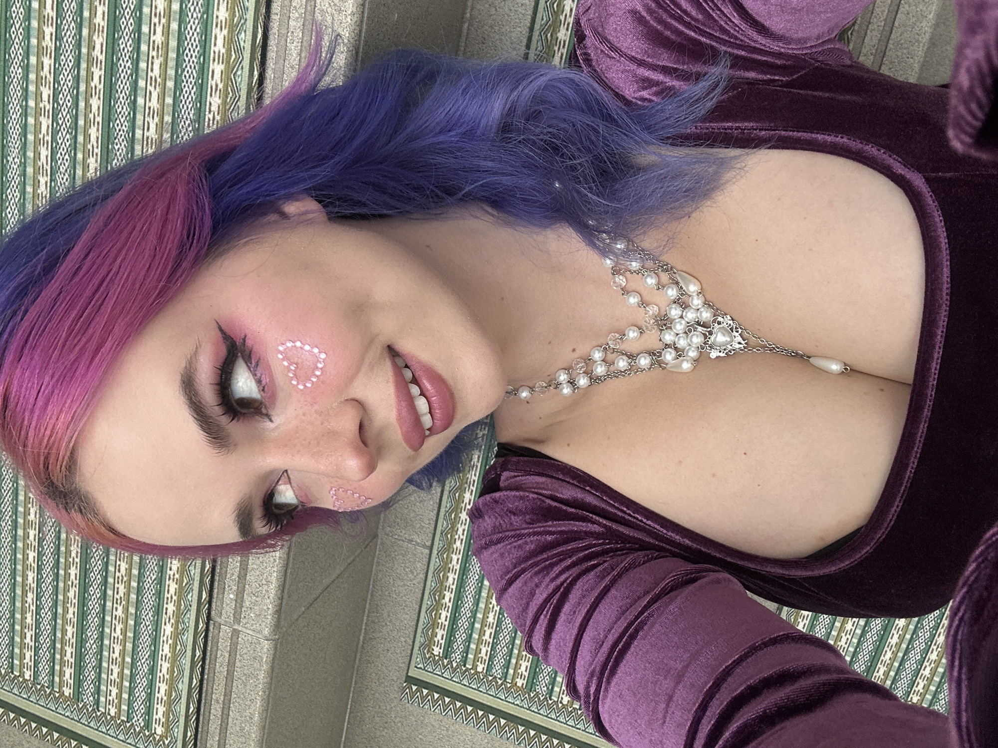 A woman with pink and purple hair, wearing a purple velvet dress and a pearl necklace, is lying on a rug with a geometric pattern. She has heart-shaped decorations on her face. The rug is on a light gray tile floor. The image appears to be taken indoors. There is no specific information to identify the location as City of Zagreb, Croatia.