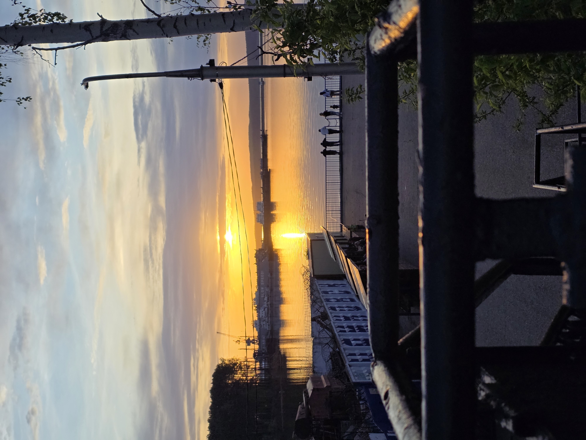 Captured from the city of Chaikovsky, Russia, the image portrays a sunset over what appears to be the Kama River. The sun's reflection dominates the water, creating a golden hue. A group of people is standing along a waterfront walkway, observing the sunset. A lamppost extends into the sky. In the foreground, a metal structure and some objects are visible, creating a sense of depth. Other structures and a crane are on the shore further down the river.