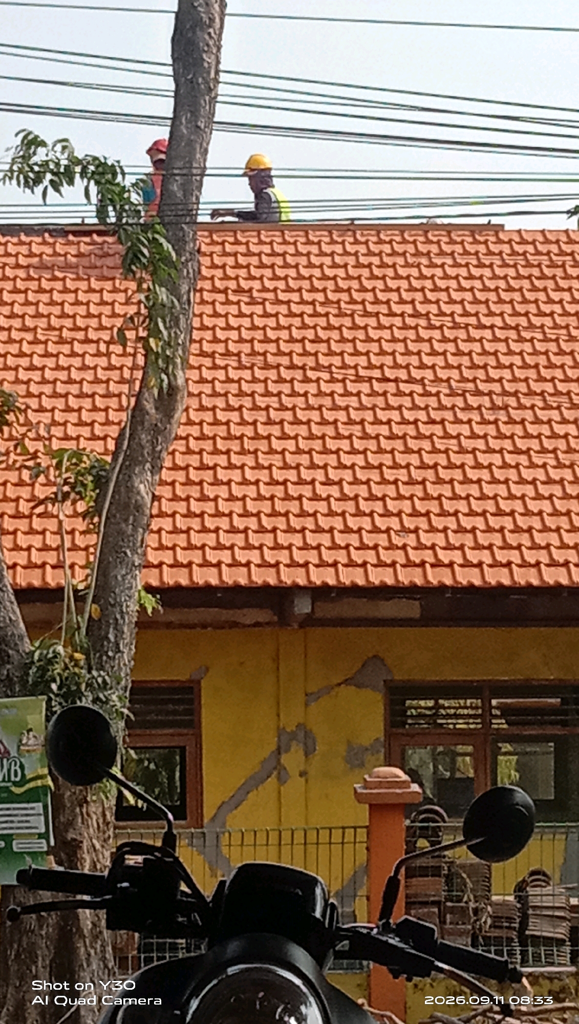 Two individuals wearing hard hats and safety vests are on the roof of a building with orange tiles. One individual, wearing a red hard hat and a blue and orange vest, is partially obscured by a tree trunk. The other individual, wearing a yellow hard hat and a yellow vest, is leaning over the roof's edge. A tree with green leaves stands to the left of the building. The building has a yellow exterior with visible cracks and two windows with wooden frames. A portion of a motorcycle is in the foreground, and utility lines are visible in the sky. The scene appears to be in the city of Munggugianti, Indonesia, given the timestamp "2026.09.11 08:33" which indicates a date format common in Indonesia and the mention of "Shot on Y30 AI Quad Camera" which is a mobile phone model.