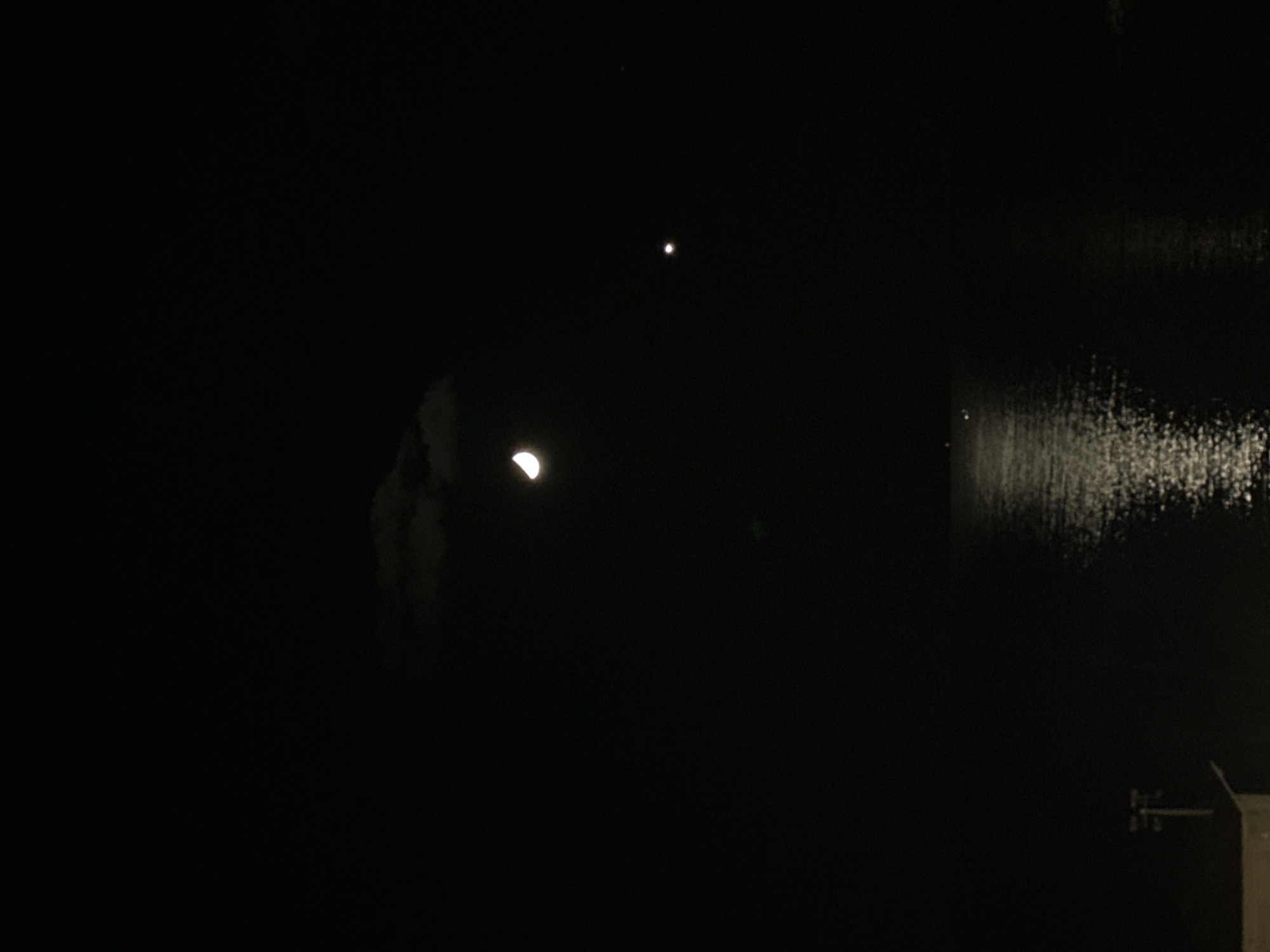 The image depicts a night scene in Tel-Aviv, Israel. A crescent moon is visible in the dark sky, along with a single bright star or planet positioned above it. Reflections of light, possibly from city lights, are seen on a body of water to the right of the image. There are some dark structures visible in the bottom right corner, potentially parts of buildings or infrastructure.