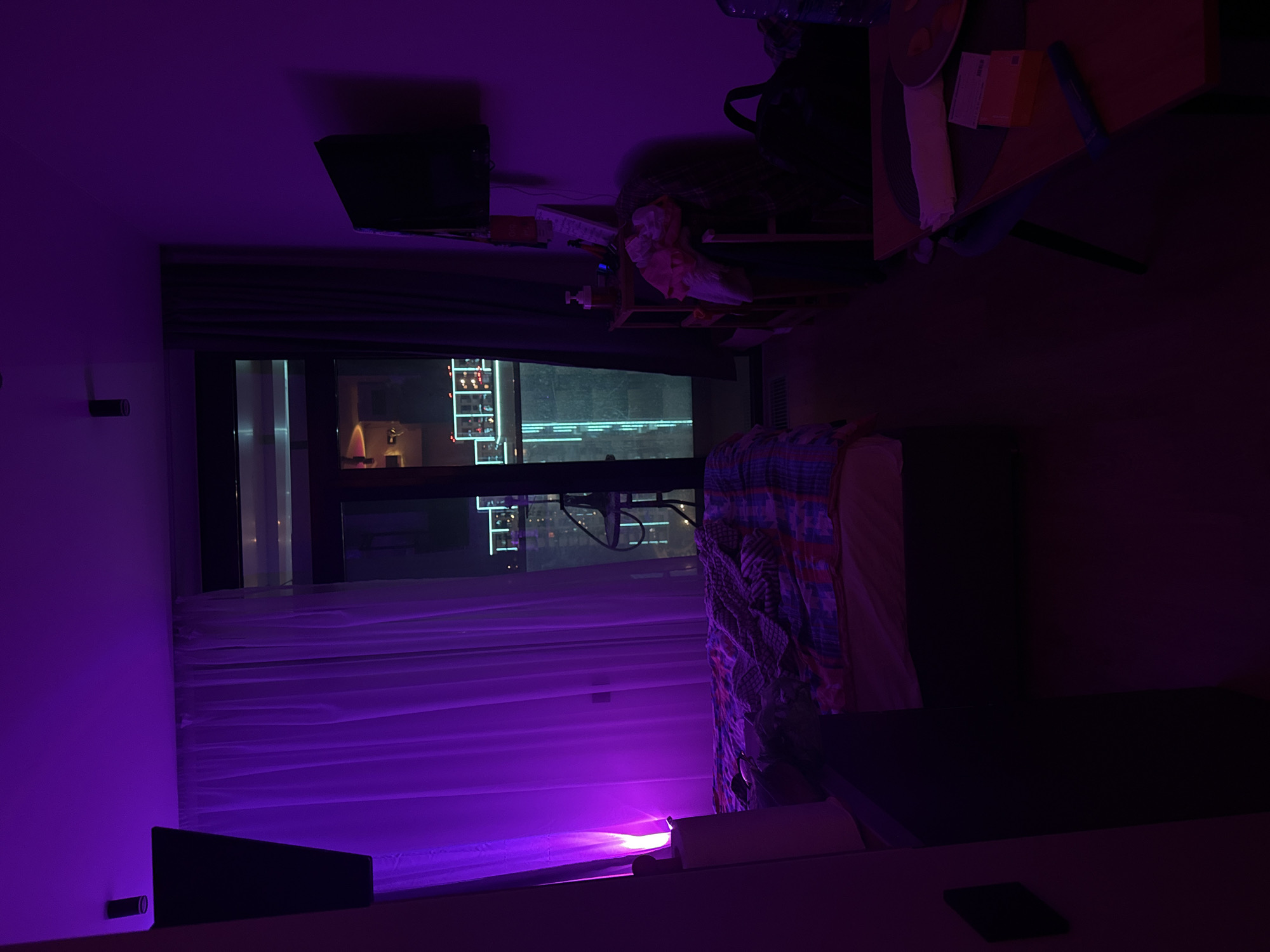 The image shows the interior of a room in Sochi, Russia, at night. A window offers a view of the city lights outside. The room is illuminated by a purple light source. A bed with patterned bedding is visible, along with a nightstand with various items on it, including papers and what appears to be a beverage bottle. A television screen is mounted on the wall near the window. The room is generally dark and somewhat cluttered. There are no people visible in the image. A small device, possibly a phone or tablet, is on a surface near the bottom of the frame.