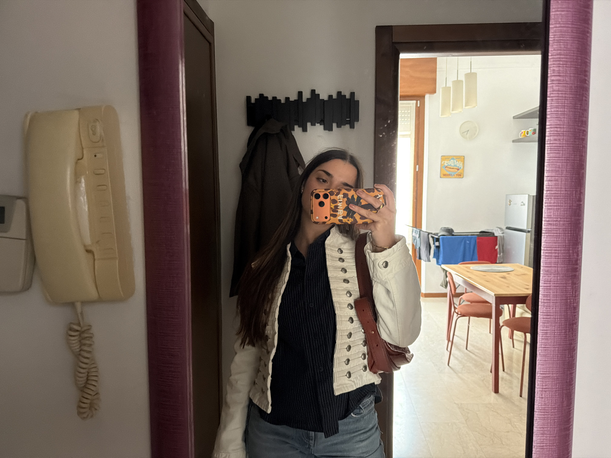 The image shows a person reflected in a mirror, taking a photo of themselves. The person has long brown hair, wears a black shirt, a white jacket, and blue denim shorts, and is holding a smartphone with a patterned orange and brown case. A brown shoulder bag hangs from the person's shoulder.
The background, visible through the mirror, reveals a room with a dining table and chairs, clothes hanging to dry, and kitchen appliances. A coat rack with a brown coat is seen behind the person. A wall-mounted phone is to the left of the mirror. The image was taken in Milan, Italy based on the design and appearance of the interior.
