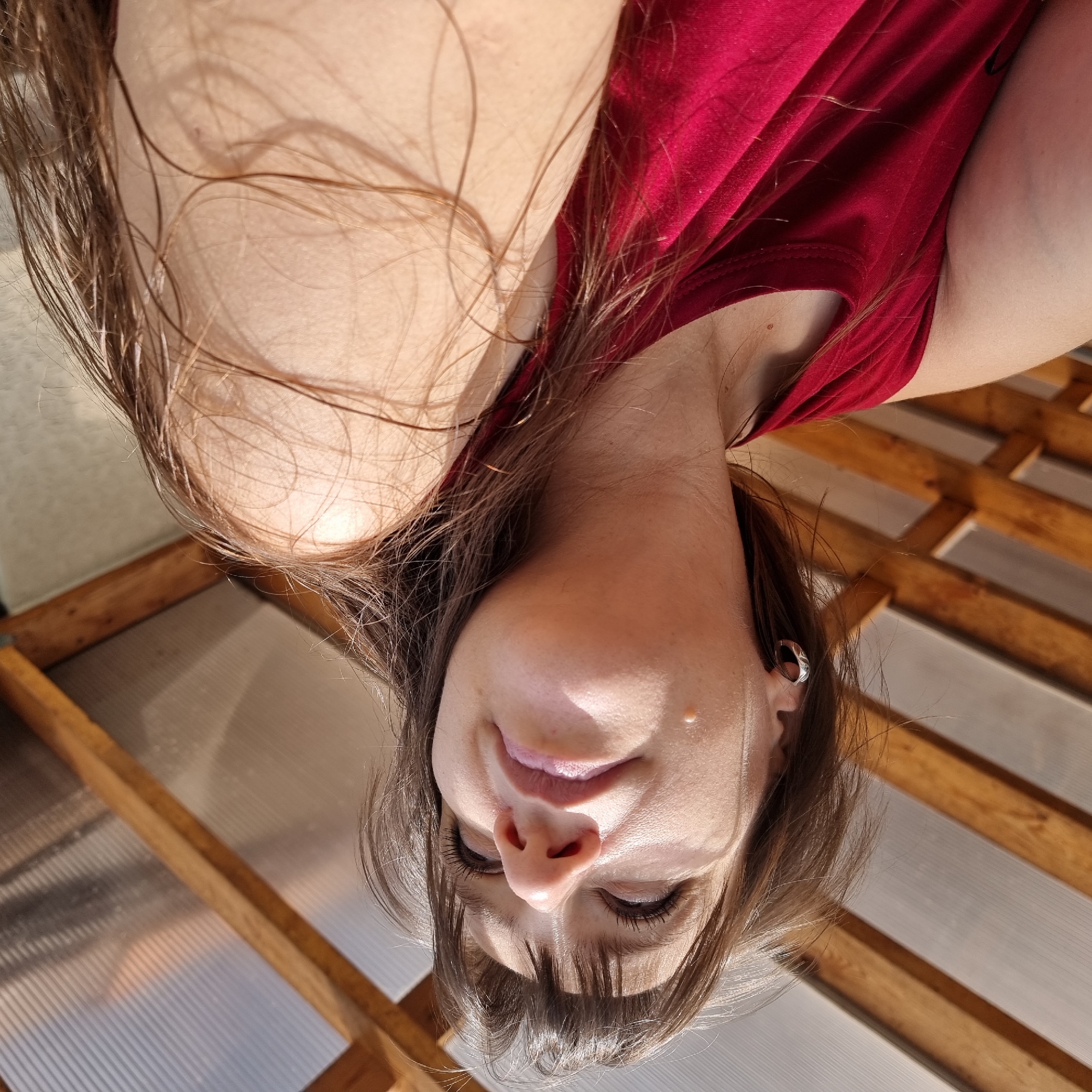 A low-angle, medium shot captures a fair-skinned person with long, brown hair and bangs, looking upwards and to the right. The person is wearing a sleeveless, red top. Their left ear is visible, adorned with a silver hoop earring. A small, light-colored mole is noticeable on their cheek. The background consists of a wooden and translucent-roofed structure, possibly a patio or greenhouse, under a bright, sunny sky. The lighting suggests it is daytime. The image does not contain any elements that would allow for a specific location in Dunnae-myeon, South Korea to be determined.