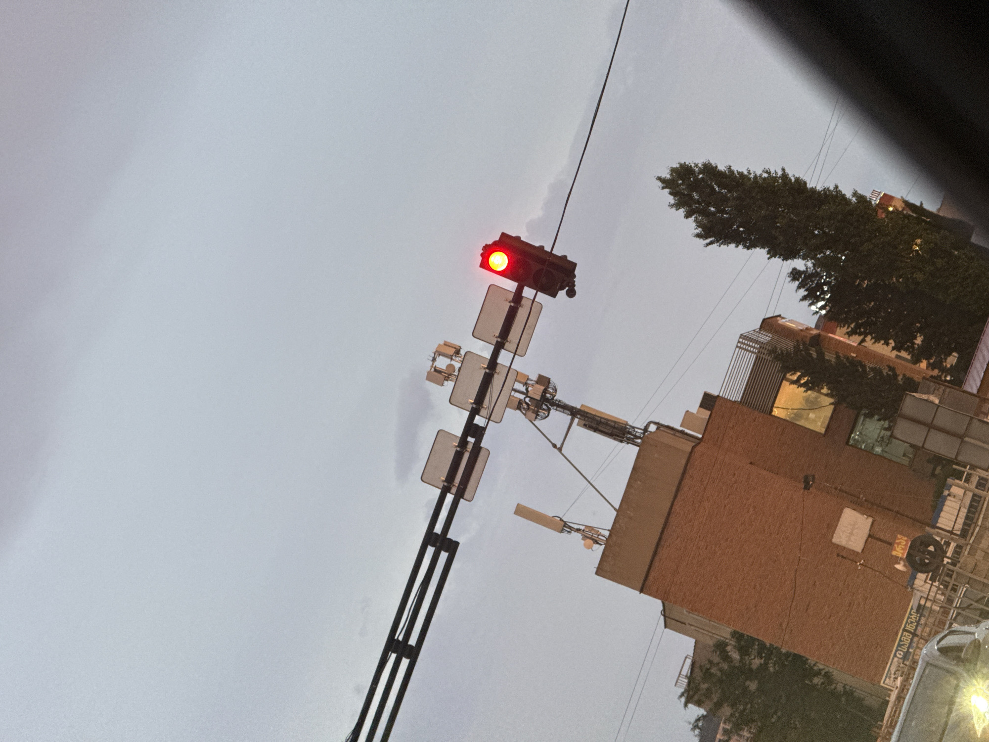 This media file shows a street scene in Tbilisi, Georgia, with a traffic light displaying a red signal. The traffic light is mounted on a pole along with several signs. In the background, there are buildings, some of which appear to be under construction or renovation, with exposed brickwork. Trees are also visible, with lush green foliage. The sky is overcast, suggesting it might be a cloudy day or dusk. The overall scene is shot from a low angle, looking upwards. There are no people visible in the frame, and the focus is on the infrastructure and the urban environment. The image has a slightly tilted perspective, adding a dynamic element to the composition.