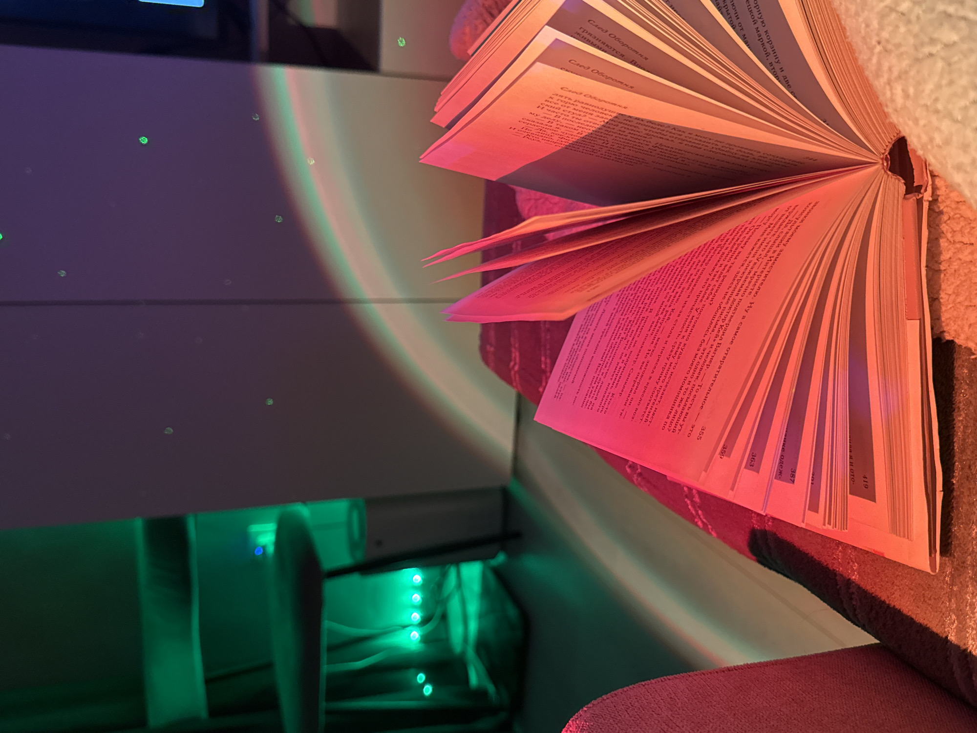Here is a matter-of-fact description of the image:
The image shows an open book placed on a pink surface, possibly a cushion or fabric. The book is oriented vertically, with its pages spread out. The pages are filled with text, appearing to be in a language other than English. There are page numbers visible, indicating sections or chapters within the book.
In the background, there is a white surface with small dots, possibly reflections or decorative elements. To the left, there is a dark, possibly metallic or plastic structure with green LED lights emanating from within it. The lighting in the image appears to be artificial. The overall arrangement suggests an indoor setting, possibly a room with various light sources and decorative elements.