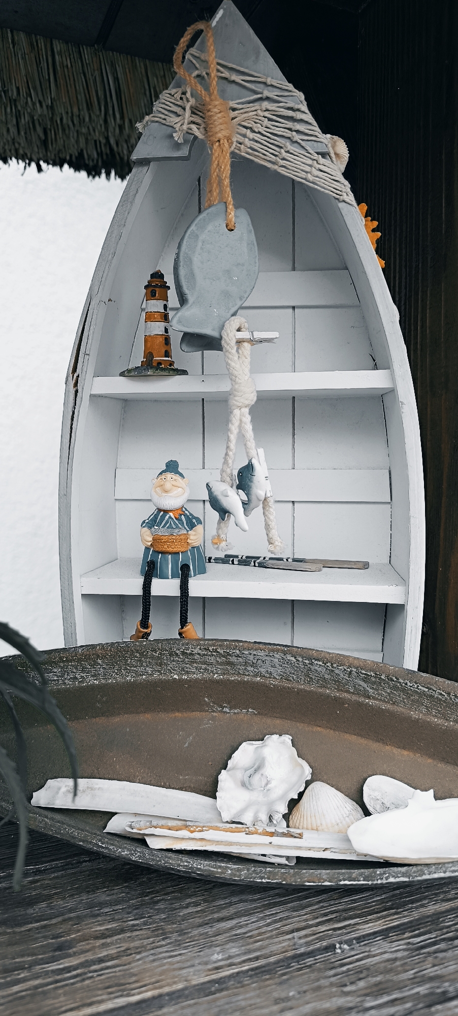 The image shows a decorative display with a nautical theme, likely set up outdoors. The central subject is a white, shelf-lined structure shaped like the hull of a boat. On the top shelf, a miniature lighthouse and a grey, oval object hang from a rope attached to a macrame net. A small, smiling figurine of a man with a beard and a striped shirt sits on the second shelf, holding a basket. Two small fish figurines are suspended from a rope below him. On the shelf below, two small wooden sticks with black and white stripes are placed. The boat structure rests on a curved, dark grey surface, which in turn sits on a rustic wooden table. The surface beneath the boat contains several white seashells and bone-like fragments. The background is a soft, blurry white on the left and a dark, textured surface on the right, possibly wood. The overall impression is of a charming, handcrafted display evoking a seaside or fishing village aesthetic. The lighting suggests it is daytime.