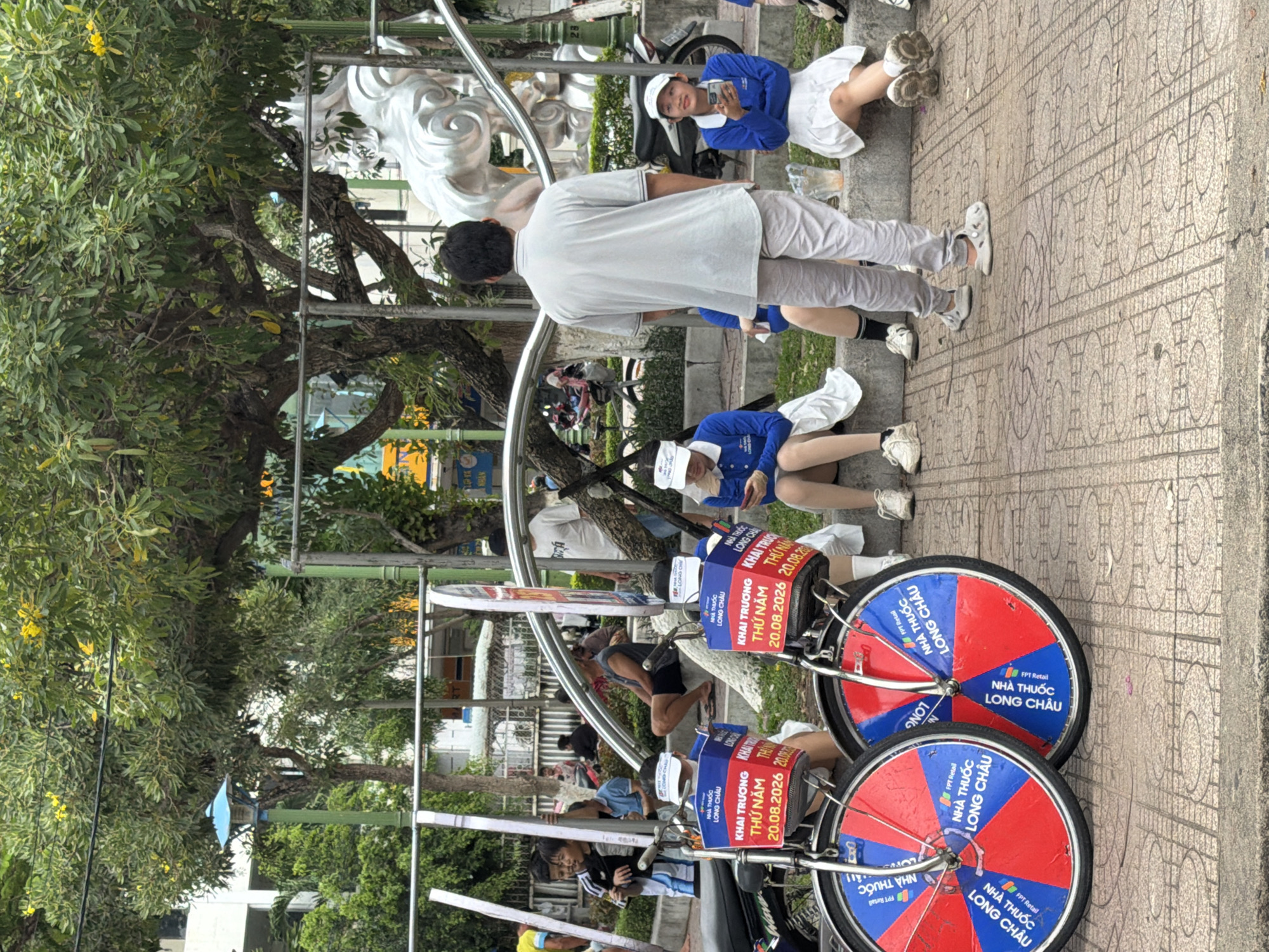 This image depicts an outdoor scene in Nha Trang, Vietnam, likely in a park or public square. In the foreground, two bicycles with red and blue wheels are parked on a tiled pavement. The wheels feature text in Vietnamese, including "NHÀ THUỐC LONG CHÂU" and "FPT Retail". A sign on the handlebars of the bicycles reads "KHAI TRƯƠNG THỨ NĂM 20.08.2026". Several people are seated on a low concrete ledge behind the bicycles. Some are wearing blue shirts and white skirts, and at least one is holding a mobile phone. A man in a grey t-shirt and light-colored pants stands facing away from the camera, looking towards the background. Further back, a large decorative sculpture of a horse is visible. Trees with green foliage and some yellow flowers provide shade, and a building can be seen in the upper left corner. The overall atmosphere suggests a public event or gathering.