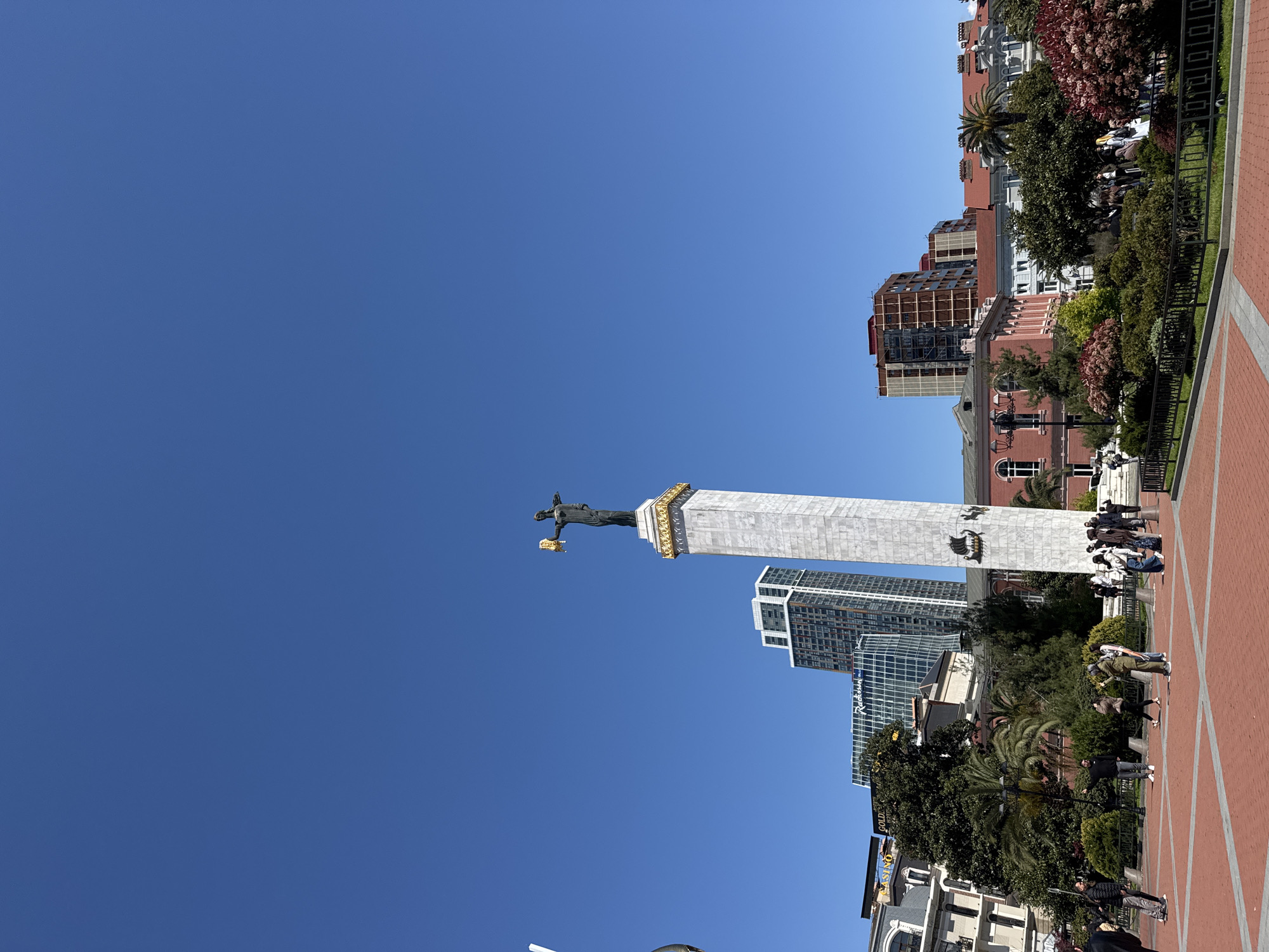 The image shows the Statue of Liberty in Batumi, Georgia. The statue stands atop a tall, white column adorned with gold trim. The sky is a clear, bright blue. Surrounding the statue at ground level is a brick-paved plaza with trees and other greenery. Buildings, including modern high-rises and older, red-roofed structures, can be seen in the background. Several people are walking around the plaza.