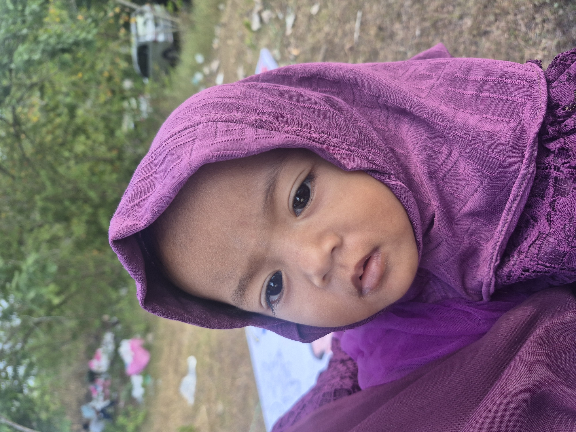 A close-up portrait shows a baby, likely a girl, looking directly at the camera with a neutral and observant expression. The baby is wearing a vibrant purple head covering, possibly a hijab or scarf, which features a distinctive textured pattern of small geometric shapes. Her dark eyes are wide and focused. She is dressed in matching purple attire; details on the right side of the frame reveal sections of sheer and patterned fabric, suggesting a layered garment. The background is out of focus, featuring green foliage from trees and bushes, indicating an outdoor setting. On the ground behind the baby, blurry shapes, possibly discarded items like plastic or clothing, are visible. A white vehicle is partially discernible in the upper left background. The natural lighting suggests it is daytime. No discernible text is visible. The scene is set outdoors, likely in a casual environment in Lhokseumawe, Indonesia.