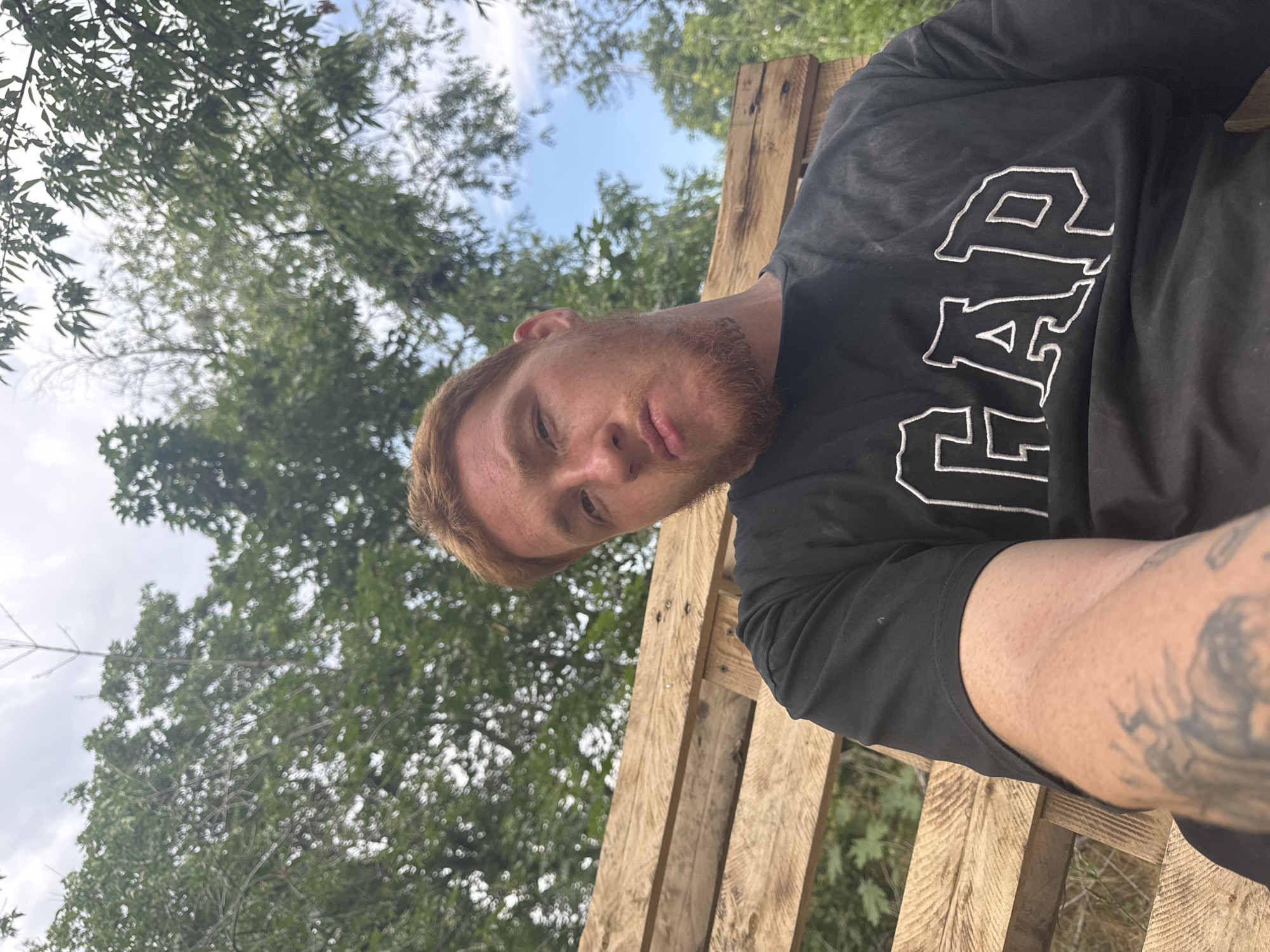 A man with red hair and a beard is sitting on a wooden structure made of pallets. He is wearing a black t-shirt with "GAP" written on it in white letters. The background consists of trees and a cloudy sky. He is looking directly at the camera. The image appears to have been taken outdoors. There is a tattoo on his left arm.