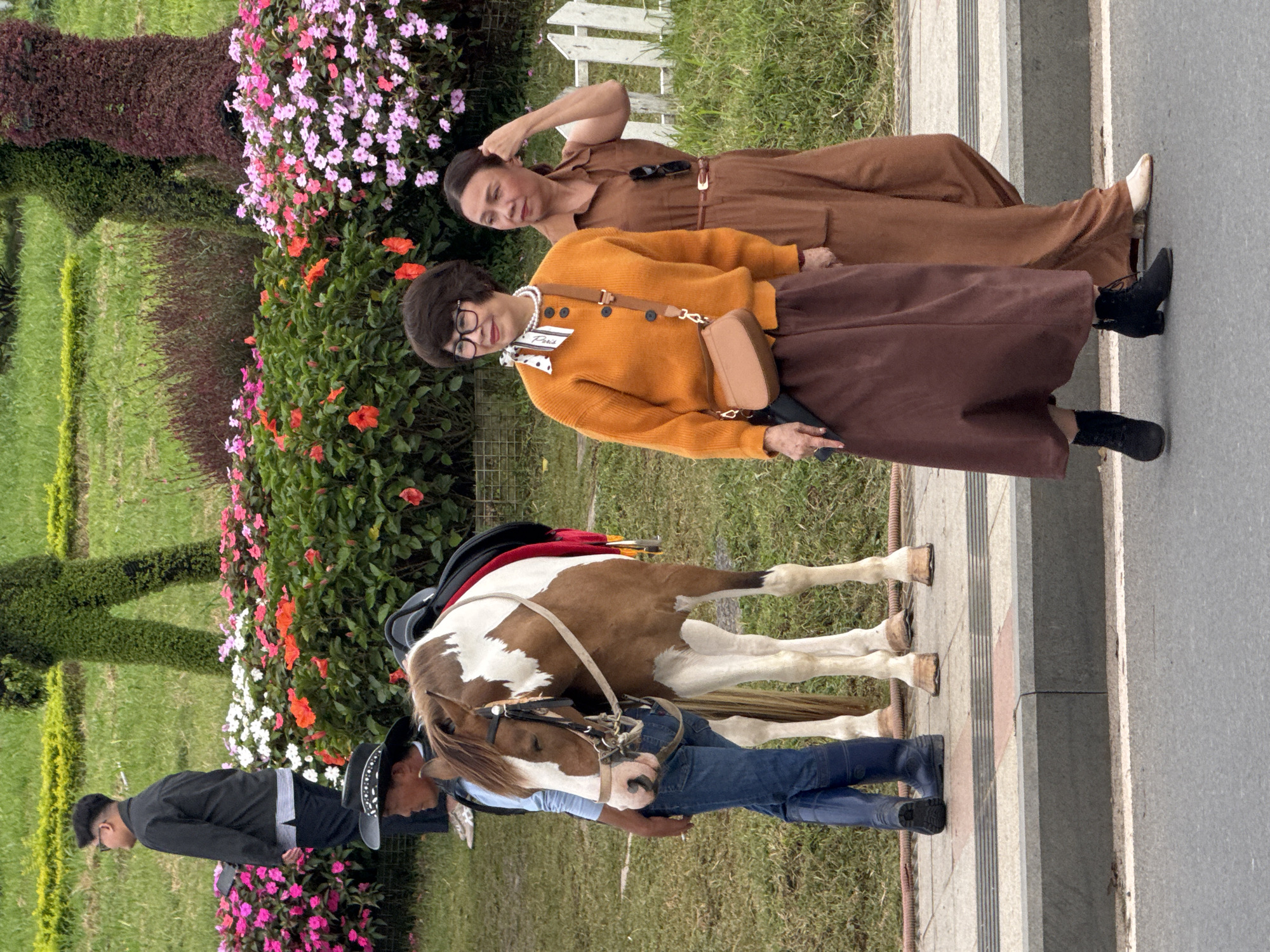 This image depicts a scene in the city of Da Lat, Vietnam, likely at a park or garden area. In the foreground, two women are walking on a paved pathway next to a road. The woman on the left is wearing an orange cardigan over a brown skirt and black boots, with a brown crossbody bag. The woman on the right is wearing a brown jumpsuit and white shoes. They are walking towards the right side of the frame. Behind them, a man in a cowboy hat and blue jeans stands next to a brown and white horse. The horse is saddled and appears to be waiting. In the background, there are colorful flower beds with pink, white, and orange blossoms, and lush green grass. Topiary figures, one resembling a person and another a pillar, are visible in the distance, adding to the garden aesthetic. The overall impression is a tranquil outdoor setting, possibly a tourist attraction, with elements of nature and human activity.