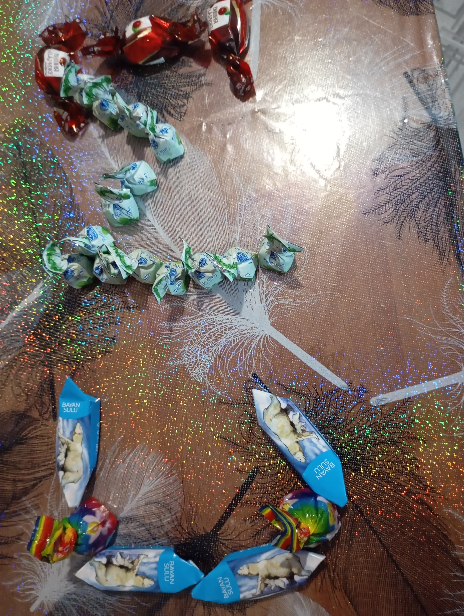 The image shows a collection of candies scattered on a textured, brown surface with a holographic, glittery pattern. The candies are wrapped in various colorful foils. Some candies are wrapped in red and gold foil, while others are in light blue and green foil with intricate patterns. A few candies have visible text or images on their wrappers, including one that appears to have an illustration of a polar bear and text that reads "BAYAN SULU". There are also a few candies wrapped in a rainbow-colored foil. The arrangement of the candies suggests they have been placed or dropped randomly on the surface. The background features a subtle pattern resembling tree branches or feathers. The lighting in the image is uneven, with a bright, washed-out area towards the top right, possibly indicating a light source. There are no people or animals visible in the scene. The context of the city of Ryabki, Russia, is not depicted in the image itself; this information is external to the visual content.