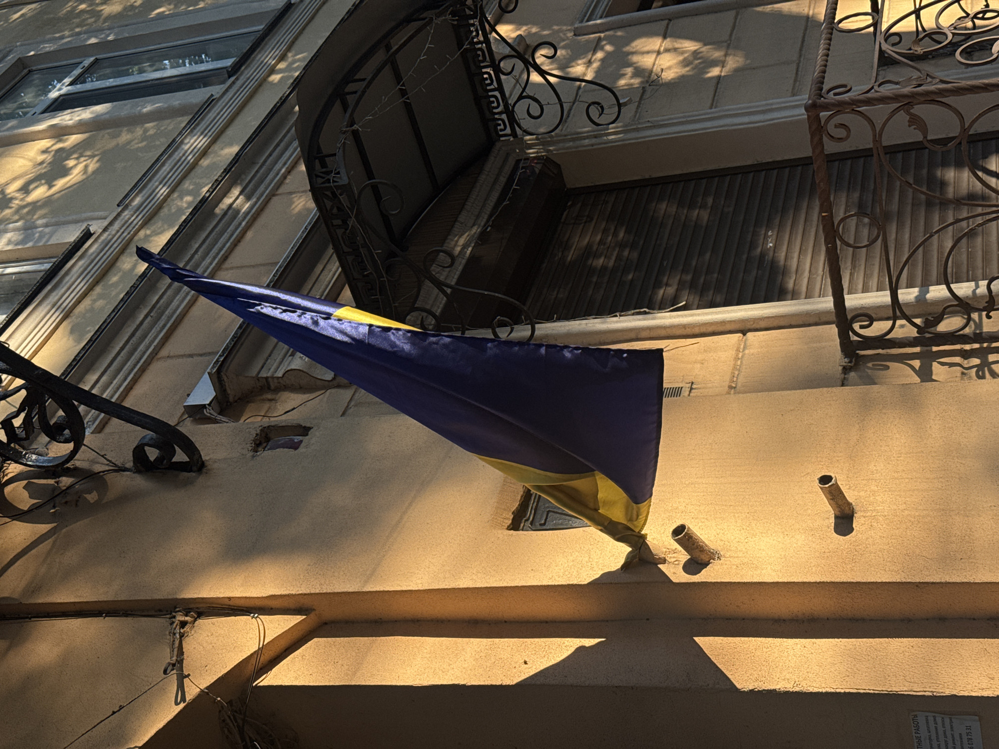 This image shows a Ukrainian flag hanging from a building in Odesa, Ukraine. The flag, with its blue and yellow colors, is draped over a textured, light-colored wall. The sun is casting long shadows across the building's facade, suggesting it is either early morning or late afternoon. The architecture features decorative wrought ironwork above the flag and a window with blinds in the background. There are also some metal pipes protruding from the wall below the flag. The overall impression is of a slightly weathered urban setting, possibly residential, under a bright sky. The flag's presence implies a sense of national identity or support for Ukraine. Visible text on a small piece of paper in the bottom right corner is not legible.