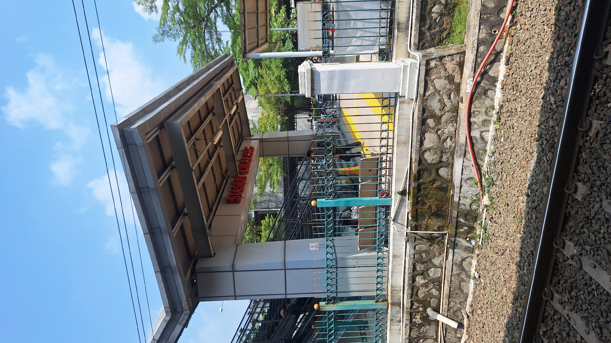The image depicts the Cikarang train station in Indonesia. A sign reading "Stasiun Cikarang" is visible. The station has a covered waiting area. The tracks are visible to the right, with a stone retaining wall and a red hose running along it. A green metal fence separates the track area from the station platform. Trees and a blue sky are in the background. People are waiting for the train.