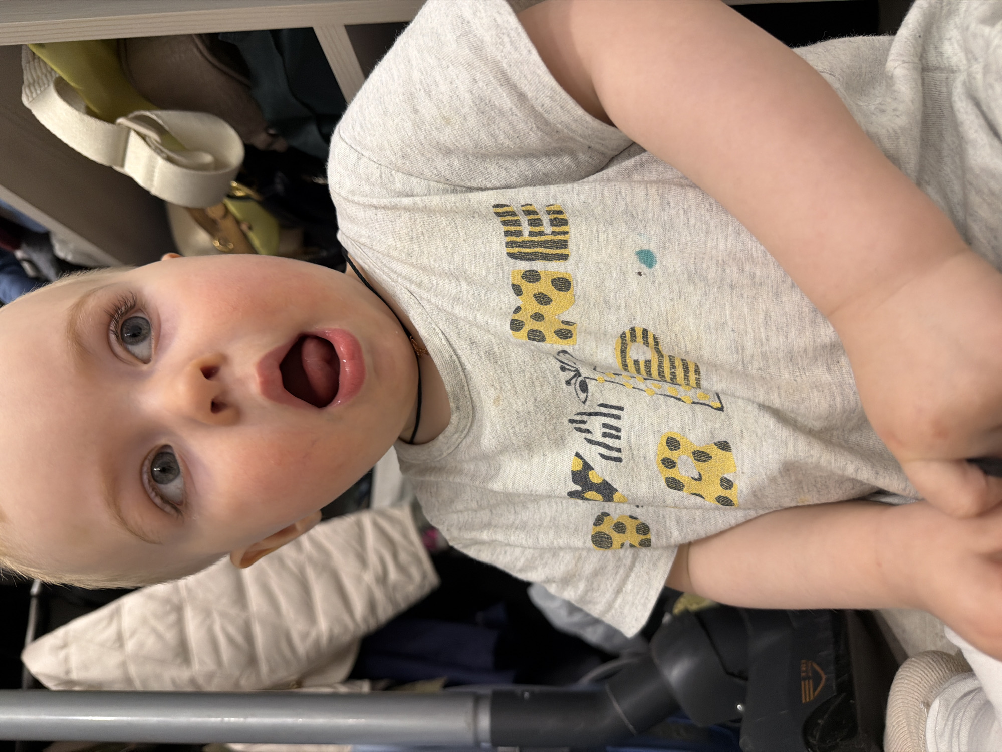 A baby with blonde hair and blue eyes is lying on its back in a closet. The baby is wearing a gray t-shirt with a yellow and black animal print design. The baby's mouth is open. Behind the baby are clothes and shelves. The location is unknown.