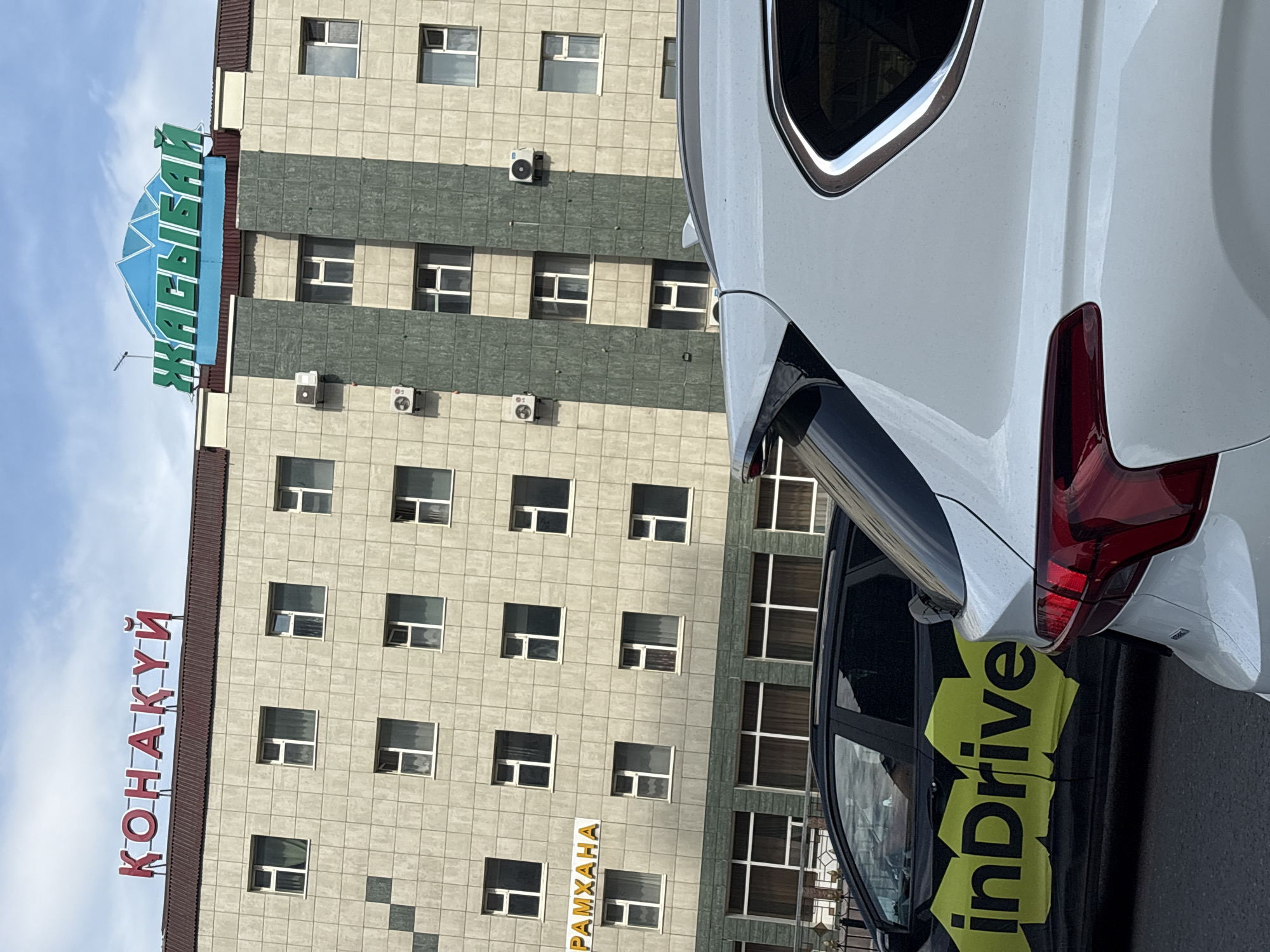 The image shows a building in Astana, Kazakhstan, with two cars in the foreground. The building has a facade of light-colored tiles and several rows of windows. There are two signs on the top of the building. The left sign, in red Cyrillic letters, reads "ҚОНАҚҰЙ". The right sign, in green Cyrillic letters and a blue triangular emblem above it, reads "ЖАСЫЛБАЙ". Below the right sign, on the facade of the building, are two air conditioning units. To the left of the building, a yellow sign with black text reads "РАМХАНА". In the foreground, a black car with yellow markings that read "inDrive" is parked next to a white SUV. The sky is cloudy.