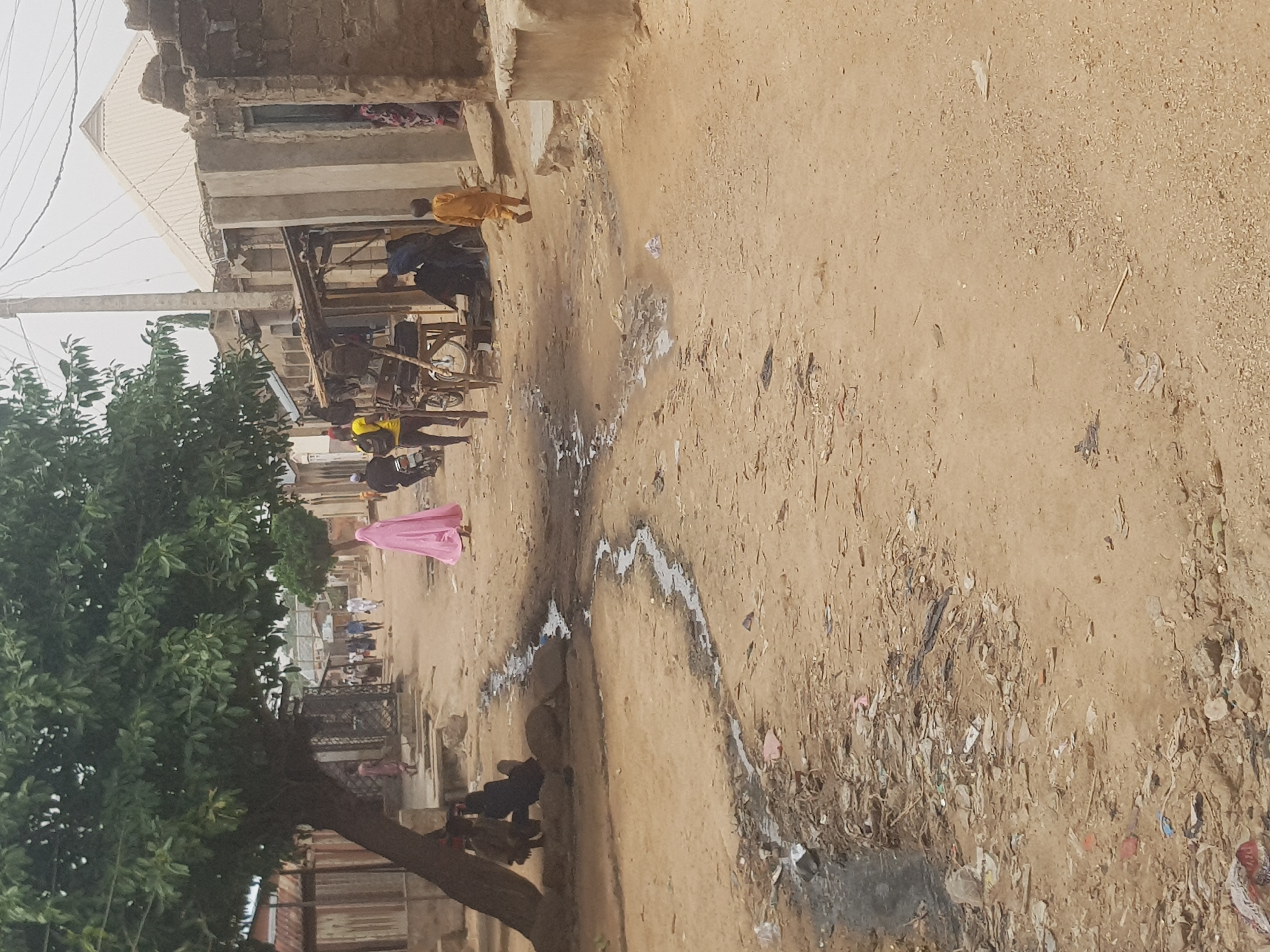 A slightly canted medium shot shows a dusty street scene in Dutsinma B, Nigeria. The dirt road is heavily littered with trash and debris.
A pink-clad figure is walking towards the center of the frame, with a group of people gathered in the distance behind the pink figure. Further up the road on the left, there are buildings and structures, and a tree. People are gathered around what appears to be a cart or stall.
