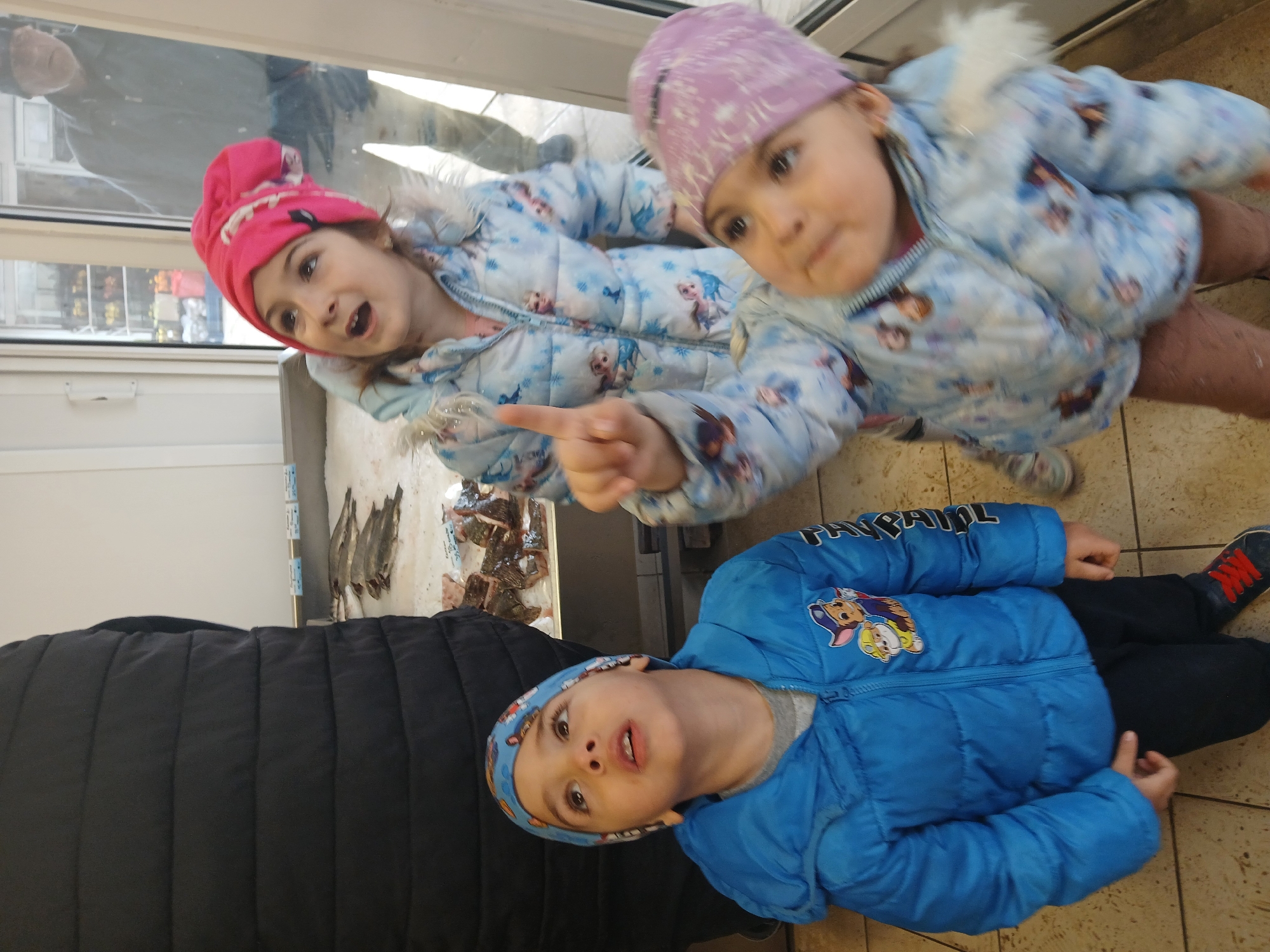 A tilted overhead shot depicts three young children indoors, possibly within or near a store in Kavarna, Bulgaria, due to the presence of fresh fish displayed in a refrigerated case visible behind the children and the reflection in the glass. Two girls, wearing winter jackets and hats, are positioned closer to the glass case. One girl with a pink hat is pointing her finger towards the fish display. The second girl, in a purple hat, is looking up at the camera. A boy wearing a blue jacket and a matching hat sits on the tiled floor, looking up at the camera. A portion of a black puffer jacket is visible in the lower left corner of the image. The lighting is bright, suggesting the store is well-lit and it is daytime.