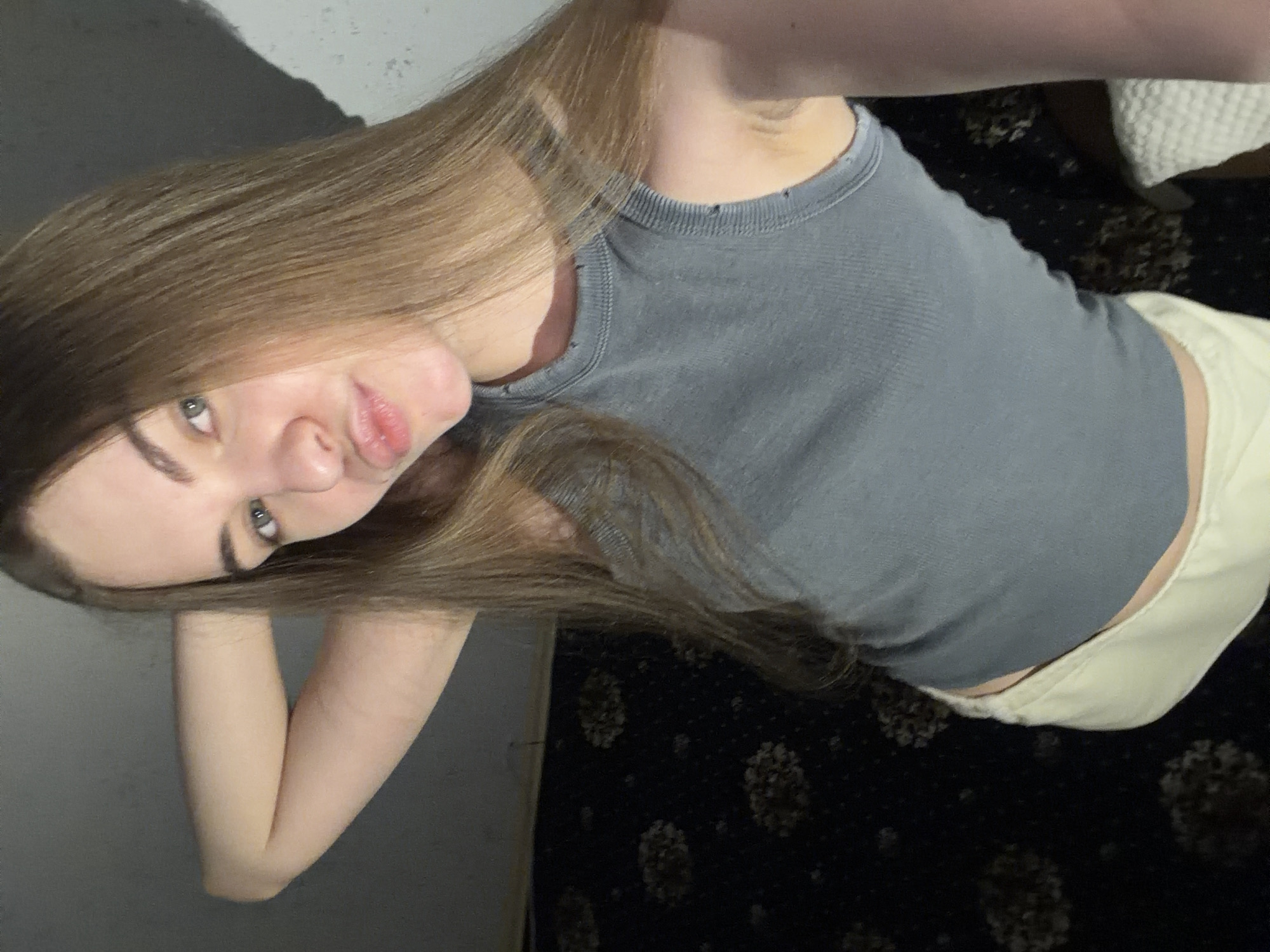 This is a media file featuring a young woman with long, light brown hair and green eyes, who appears to be in her late teens or early twenties. She is wearing a grey, ribbed tank top and light-colored shorts. The image is taken from a close-up, high-angle perspective. The woman is looking directly at the camera with a neutral expression. Her right arm is bent at the elbow and her hand is resting near her face.
The background is dimly lit and appears to be indoors. To the left of the woman, there is a grey wall with some texture. To her right, a dark fabric with a floral pattern is visible, possibly a carpet or upholstery. A white, textured object, possibly a cushion or blanket, is partially visible in the upper right corner.
The lighting suggests it might be evening or a dimly lit room. There are no clear indications of the specific time of day or weather conditions. The setting appears to be a casual, indoor environment, possibly a home. No text is visible in the image. The image has been rotated, with the woman's head appearing towards the bottom left of the frame.