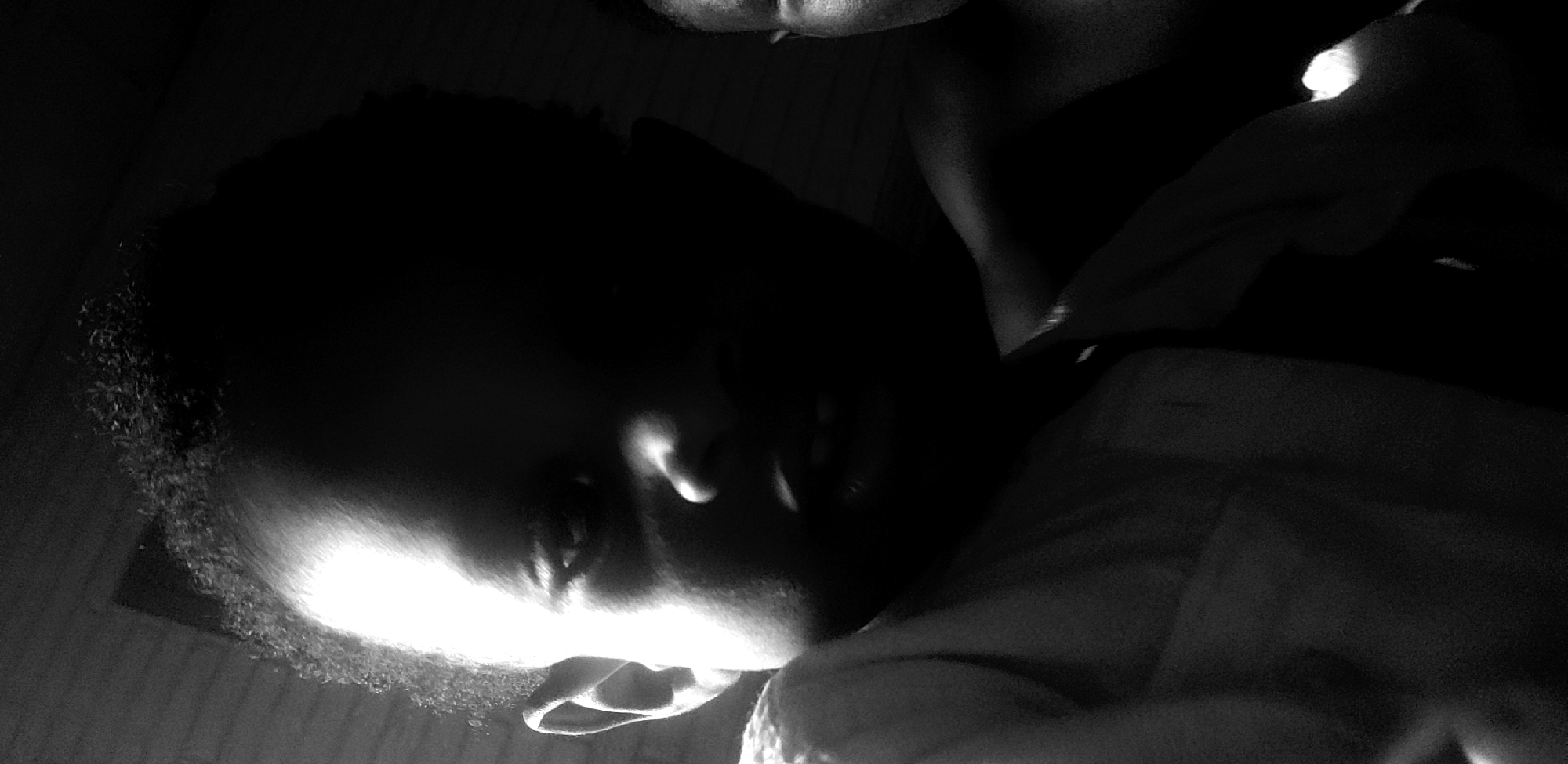 This is a black and white, close-up, low-angle shot of a person lying down in a dimly lit room, possibly a bedroom. The person, whose face is partially obscured by shadow and illuminated by a bright light source, appears to be resting or sleeping. Their head is tilted, and their facial features are softened by the lighting. The texture of their hair is visible, appearing short and curly, with some of it catching the light.
The lighting is dramatic, with strong contrasts between light and shadow, suggesting the use of a flash or a single, intense light source. The bright light highlights the contours of the person's face, particularly their cheekbone and the side of their forehead, as well as their ear. The background is dark and indistinct, with some hints of texture that could be bedding or clothing. The overall mood is intimate and somber, with a sense of stillness. The location context provided is Narayi, Nigeria, but there are no specific visual cues within the image to confirm this setting. No text is visible in the image.
