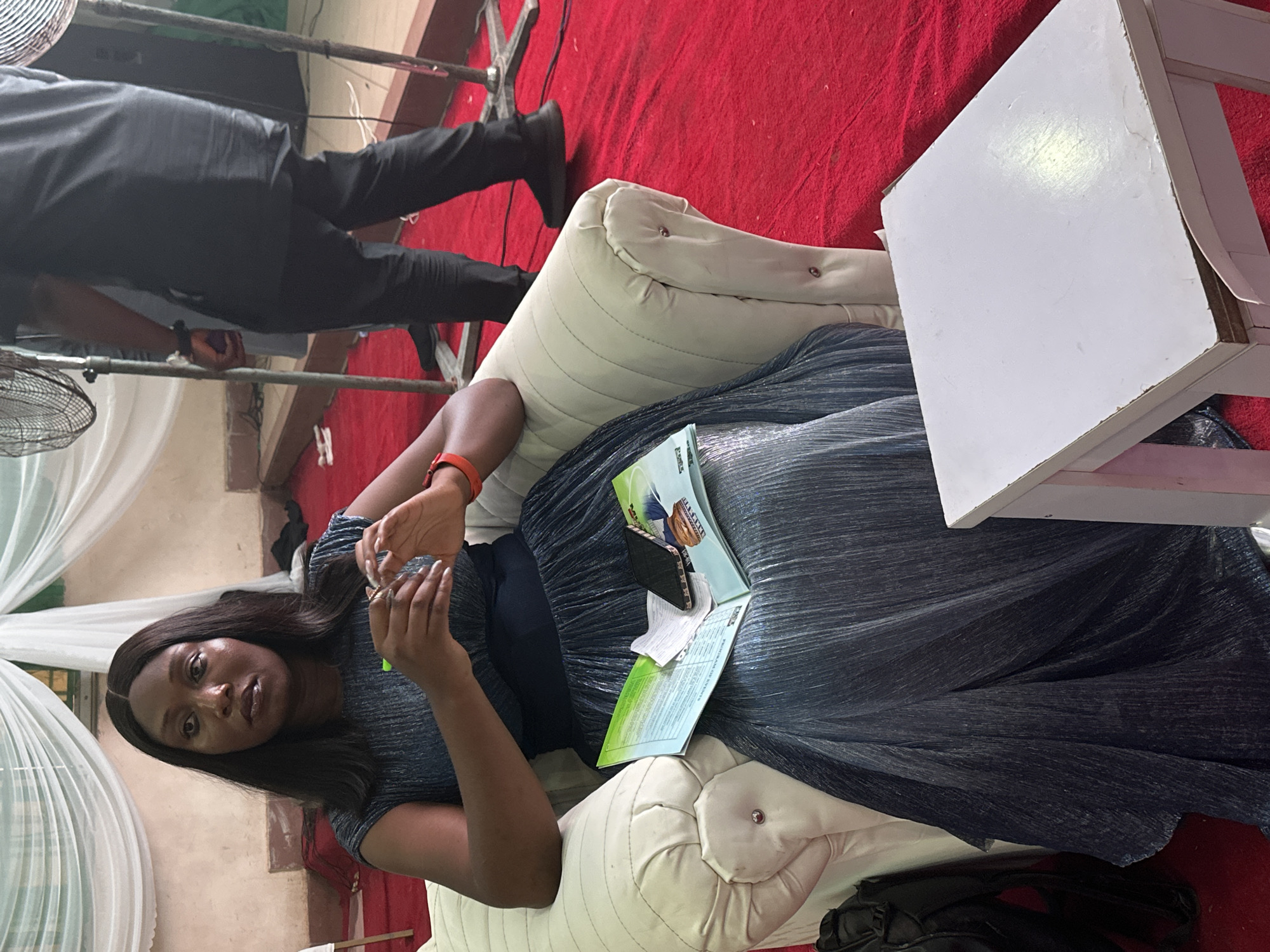 A medium shot indoors shows a woman sitting on a white sofa next to a small white table, holding something in her hands, in Unknown, Unknown. The woman has brown skin and long dark hair, and she is wearing a black dress. On top of the dress is a green and white document. A black wallet is on top of the document. A person with dark pants and black shoes is standing behind the sofa. A red carpet is behind the sofa and white table.