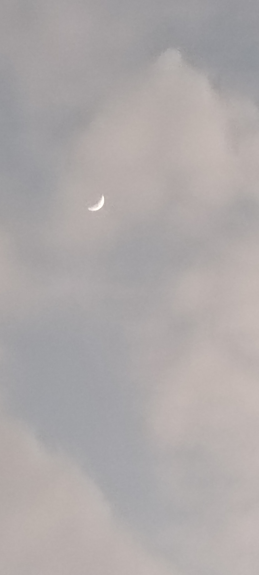 A faint crescent moon is visible in the sky above Lhokseumawe, Indonesia, partially obscured by clouds. The sky is a pale gray, with softer, lighter gray clouds dominating the frame. The moon, a sliver of white, is positioned in the upper left quadrant of the image. There are no people or man-made objects visible in the scene, creating a serene and natural landscape.