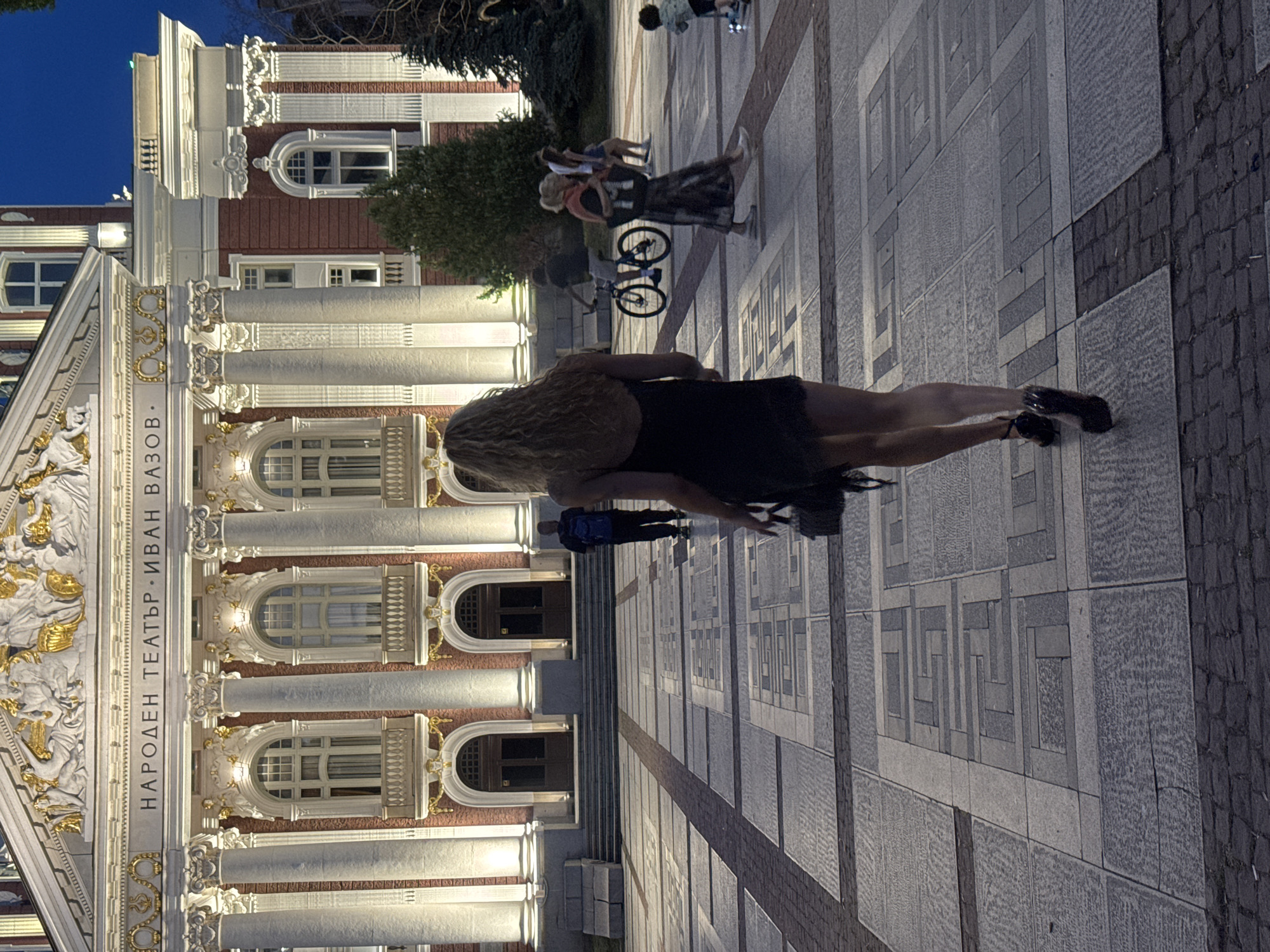 The image depicts a public square in front of the Ivan Vazov National Theatre in Sofia, Bulgaria. The theatre, an ornate neoclassical building, is illuminated by artificial lights. In the foreground, a woman with curly blonde hair walks away from the camera, wearing a black dress and high heels. She is carrying a small purse. Several other individuals are visible in the background, including a person with a blue backpack walking towards the theatre, a person riding a bicycle, and a group of people gathered further in the square. The square itself is paved with large stone tiles arranged in a geometric pattern. The sky is a deep blue, indicating it is either dusk or early evening.