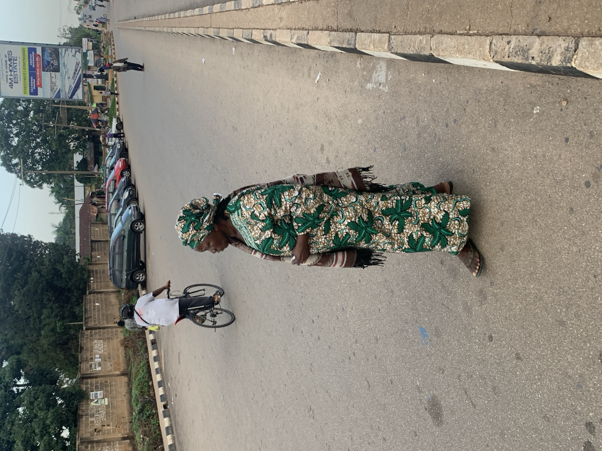 This media file shows a street scene in Barakin Lerawi, Nigeria. The primary subjects are two people: a woman in the foreground wearing a green and white patterned dress and headscarf, and a person on a bicycle in the background. The woman is walking towards the right side of the frame, with her body angled away from the camera. The cyclist, wearing a white shirt and dark pants, is also moving towards the right. Several cars are parked along the left side of the road. The setting is a paved street with a curb and sidewalk on the right. Trees and some buildings are visible in the background, as well as a large billboard. The time of day appears to be daytime, with the lighting suggesting it is not midday, possibly late morning or afternoon. The weather seems to be clear.
Visible text includes an advertisement on a billboard for "4M HOMES ESTATE" with a phone number and details about bedroom sizes and an incentive for clients. Other text is present on posters or signs on a wall in the background, but it is largely illegible. The overall atmosphere seems to be one of everyday life on a street in a Nigerian town.