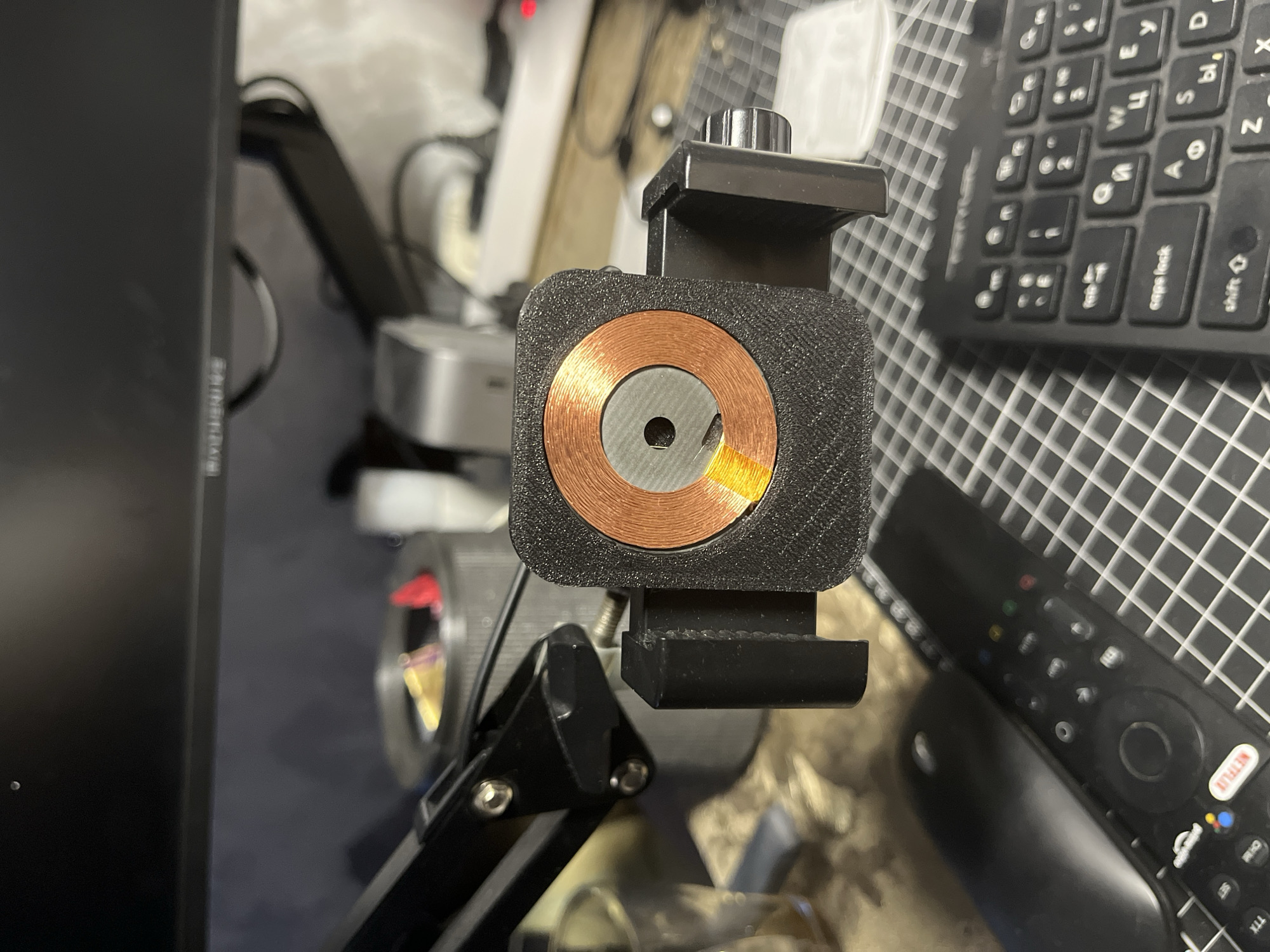 The image shows a close-up of a 3D-printed mount holding a wireless charging coil. The coil is copper-colored and has a circular design with a central grey component. The mount is black and appears to be attached to a black arm mechanism. In the background, a computer monitor is visible, along with a keyboard, a remote control, and a mouse. The overall setting appears to be a desk or workspace. There is no information in the image to determine a specific location in Moscow, Russia.