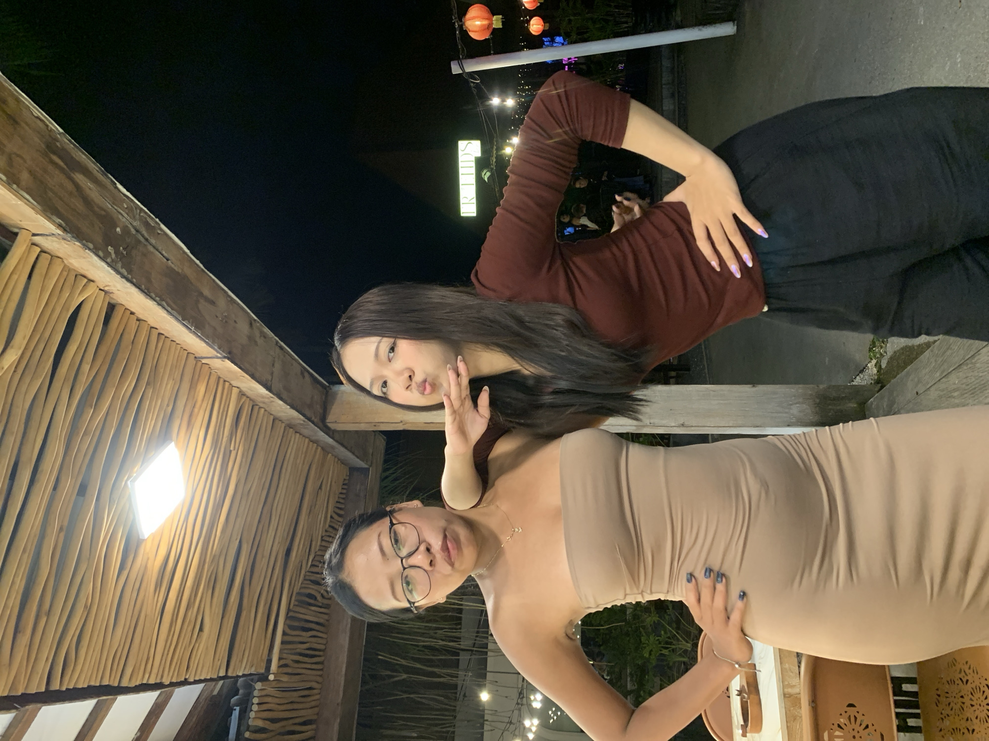 Two women are posing for a photo. One woman is wearing a brown long-sleeved shirt and black pants, while the other is wearing a beige dress and eyeglasses. The woman in the brown shirt is making a kissing face and holding up her hand. The woman in the beige dress has her hand on her hip. The background includes a wooden structure, string lights, and a sign that says "FRIENDS." The location appears to be in North Kuta, Indonesia.