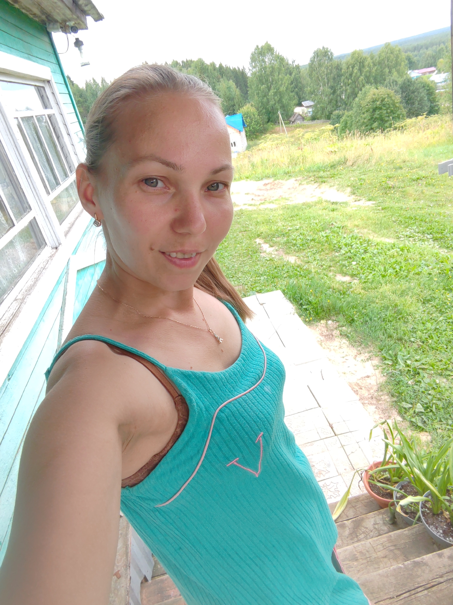 A young Caucasian woman with blonde hair pulled back in a ponytail is taking a selfie on a wooden porch in a rural area. She is wearing a turquoise tank top over a brown bra and a delicate gold necklace with a small pendant. The woman has a small mole on her left cheek and a faint smile.
Behind her, the wooden siding of a house painted teal is visible, along with a white-framed window. The porch extends to the right, with a wooden staircase leading down. In the background, there is a grassy yard with scattered yellow wildflowers and trees. Several houses and structures can be seen in the distance, nestled amongst the trees, suggesting a small village or rural settlement. The sky is overcast. The overall impression is a casual, outdoor portrait in a natural, rustic setting.