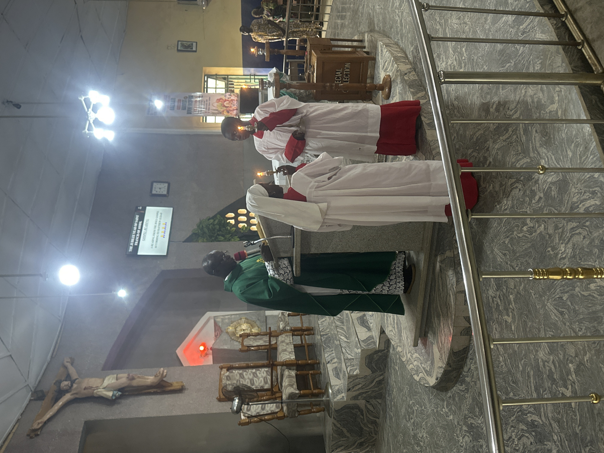 In the city of Akwanga, Nigeria, inside a church, a priest in green vestments stands at a podium, holding a microphone. Two individuals, one wearing a white robe with a red trim and the other a white head covering and robe with red trim, stand to the priest's right, holding lit candles. In the background, a crucifix is mounted on the wall, and other church furnishings are visible.