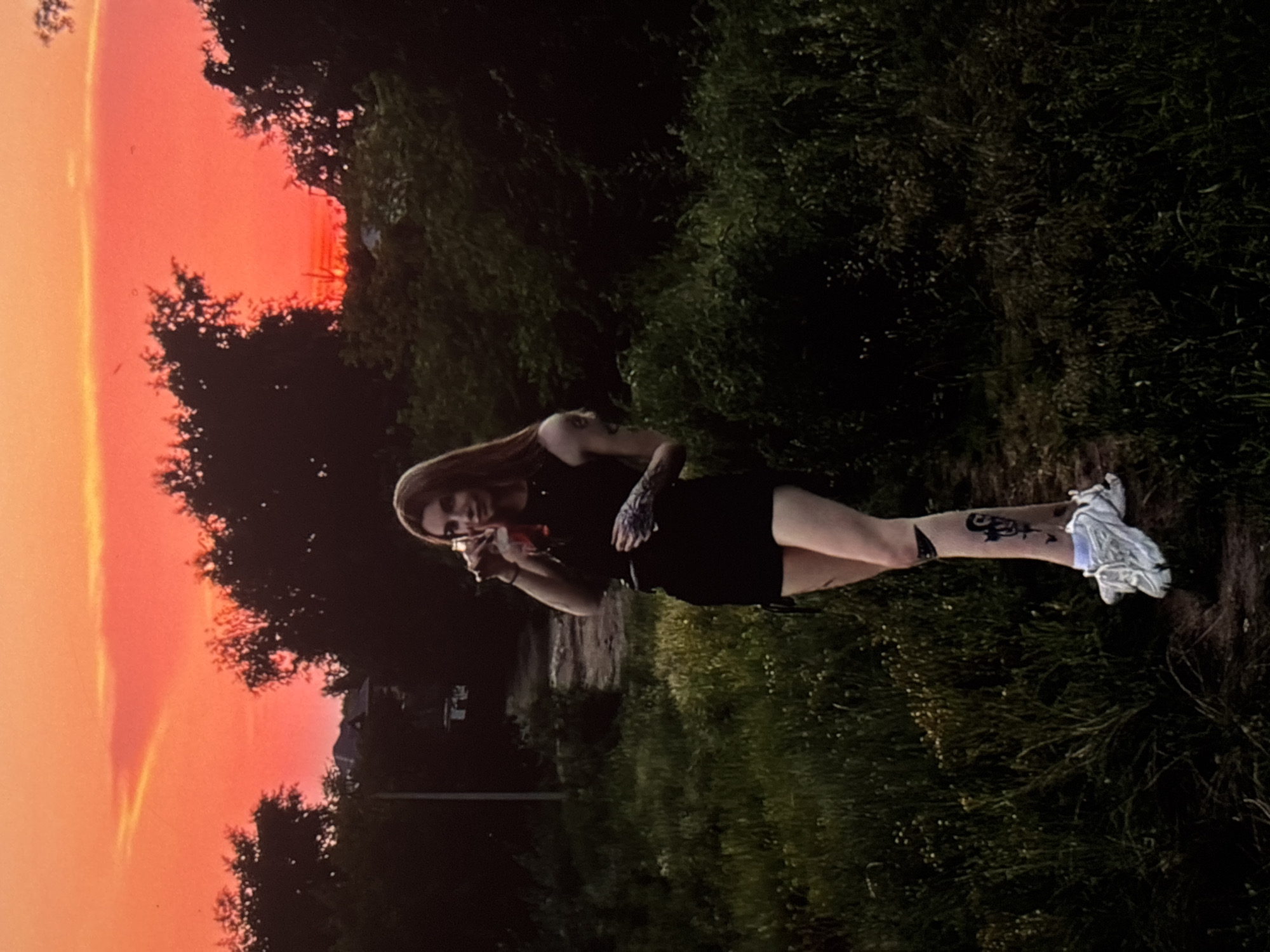 A young woman with long brown hair is standing in a field at dusk, holding a drink. She is wearing a black tank top and black shorts, with white sneakers. She has tattoos on her arms and legs. The sky is a vibrant orange and red, with silhouetted trees in the background. The image appears to be taken in a rural or semi-rural area. There is no specific information in the image to suggest it was taken in the city of Omsk, Russia.
