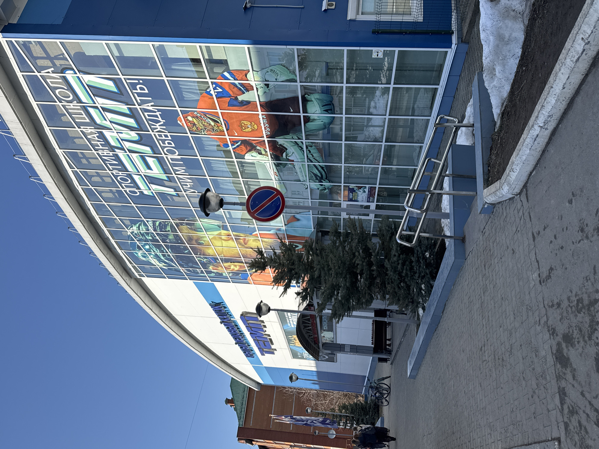 The image shows the Ice Palace "Temp" in Chaikovsky, Russia. The building is blue and white, with a large glass facade featuring an image of hockey equipment and the words "ТЕМП СПОРТИВНАЯ ШКОЛА! УЧИМ ПОБЕЖДАТЬ!". There is a "No Entry" road sign visible on the glass. A small evergreen tree grows in front of the entrance. A metal handrail and stairs lead to the entrance. The sidewalk is paved with gray bricks. A brick building with a green roof is visible to the left. There are flags and bicycles parked nearby. A few people are seen in the distance. The sky is clear and blue. Some snow patches remain on the ground.