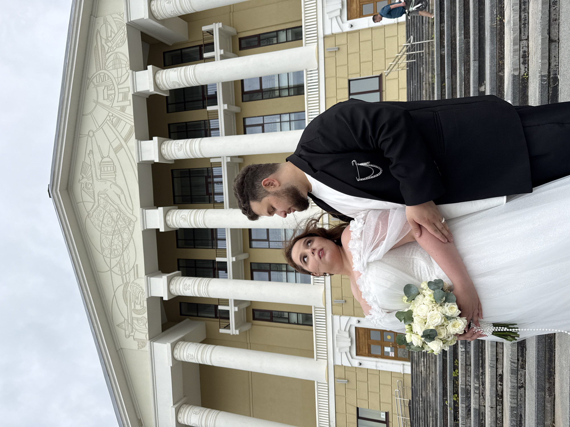 The image shows a newlywed couple posing on the steps of a building in Votkinsk, Russia. The bride is wearing a white off-the-shoulder wedding dress with floral embellishments and is holding a bouquet of white roses. The groom is wearing a black suit jacket over a white t-shirt and black pants. They are looking at each other and are positioned in the foreground. Behind them, a large building with classical architecture, featuring columns and decorative friezes, is visible. The sky is overcast. In the background, another person is walking up the stairs with a bicycle. The overall scene suggests a wedding photoshoot in front of a significant public building, possibly the Votkinsk Palace of Culture, given the architectural style.