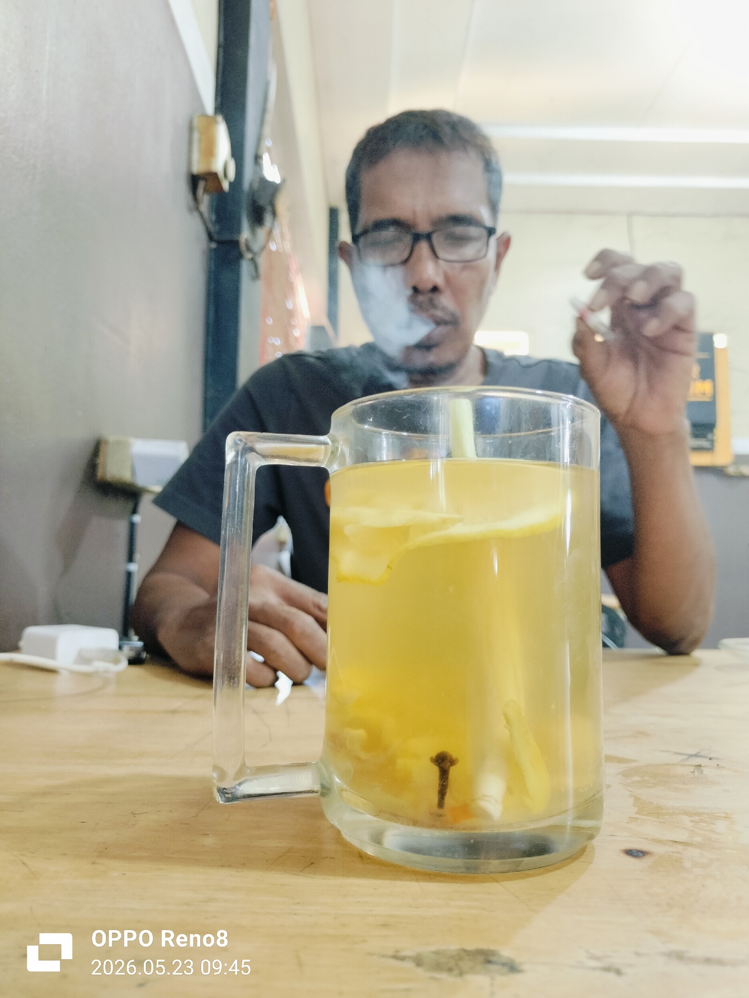 A medium, eye-level shot shows a man seated behind a wooden table with a glass mug of beverage in front of him. The man is wearing glasses and a dark gray t-shirt. He is holding a lit cigarette in his right hand, and smoke is visible near his mouth. The mug contains a yellow liquid, possibly tea or juice, with slices of fruit and what appears to be a stick of lemongrass. A white electronic device and its charger sit beside the mug. The background includes a wall, a light fixture, and partial views of the interior. At the bottom left is a watermark indicating the photo was taken with an OPPO Reno8 on May 23, 2026, at 09:45. The scene possibly takes place in Keuniree, Indonesia, based on the location.