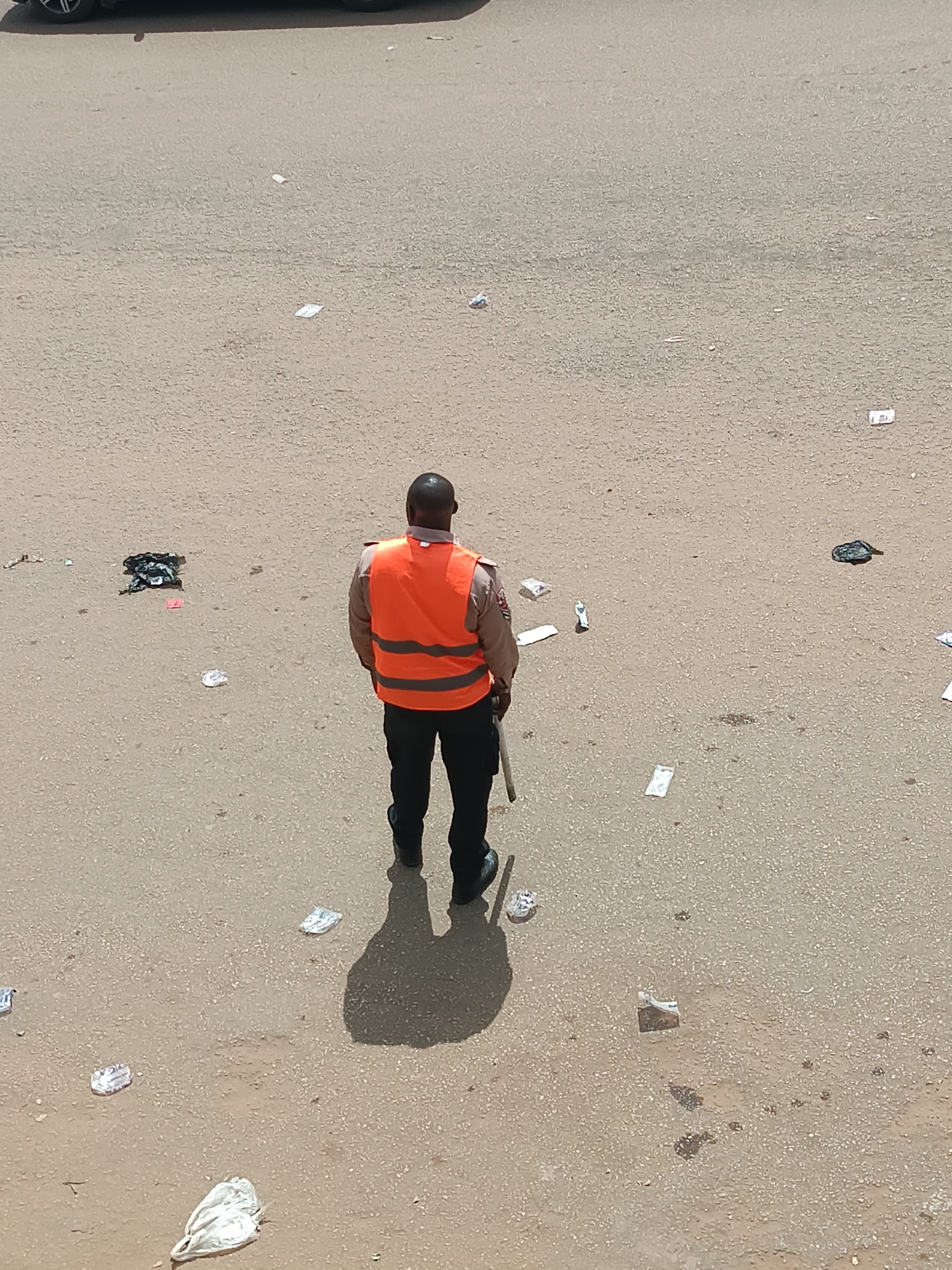 A medium-shot, overhead view shows a black man in an orange reflective vest, black pants, and black boots standing on a paved surface. He is facing away from the camera and holding a stick or baton in his right hand. The paved surface is littered with pieces of trash, including paper, plastic, and what appears to be discarded clothing. Based on context clues and available information, the image is likely taken in Bukuru, Nigeria.