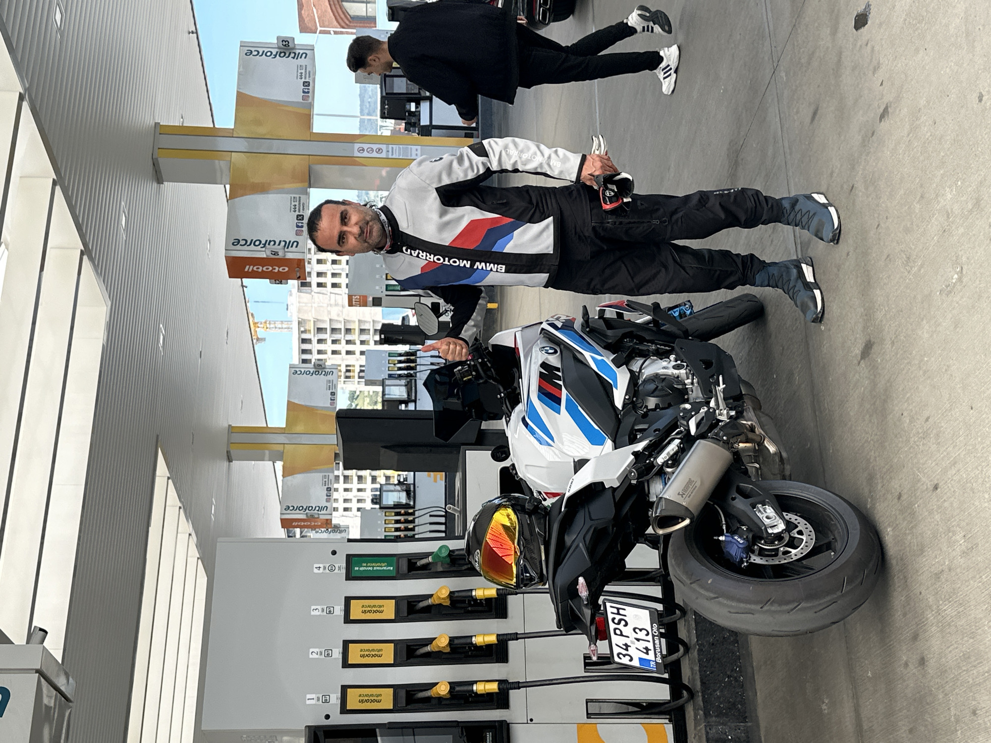 A man wearing a BMW Motorrad jacket and holding a helmet is standing beside a BMW motorcycle at a gas station. The gas station advertises "ultraforce" motorin. The motorcycle's license plate reads "34 PSH 413" with the Turkish flag. Another person is standing in the background. The photo was taken at a gas station in Gaziosmanpaşa, Turkey.