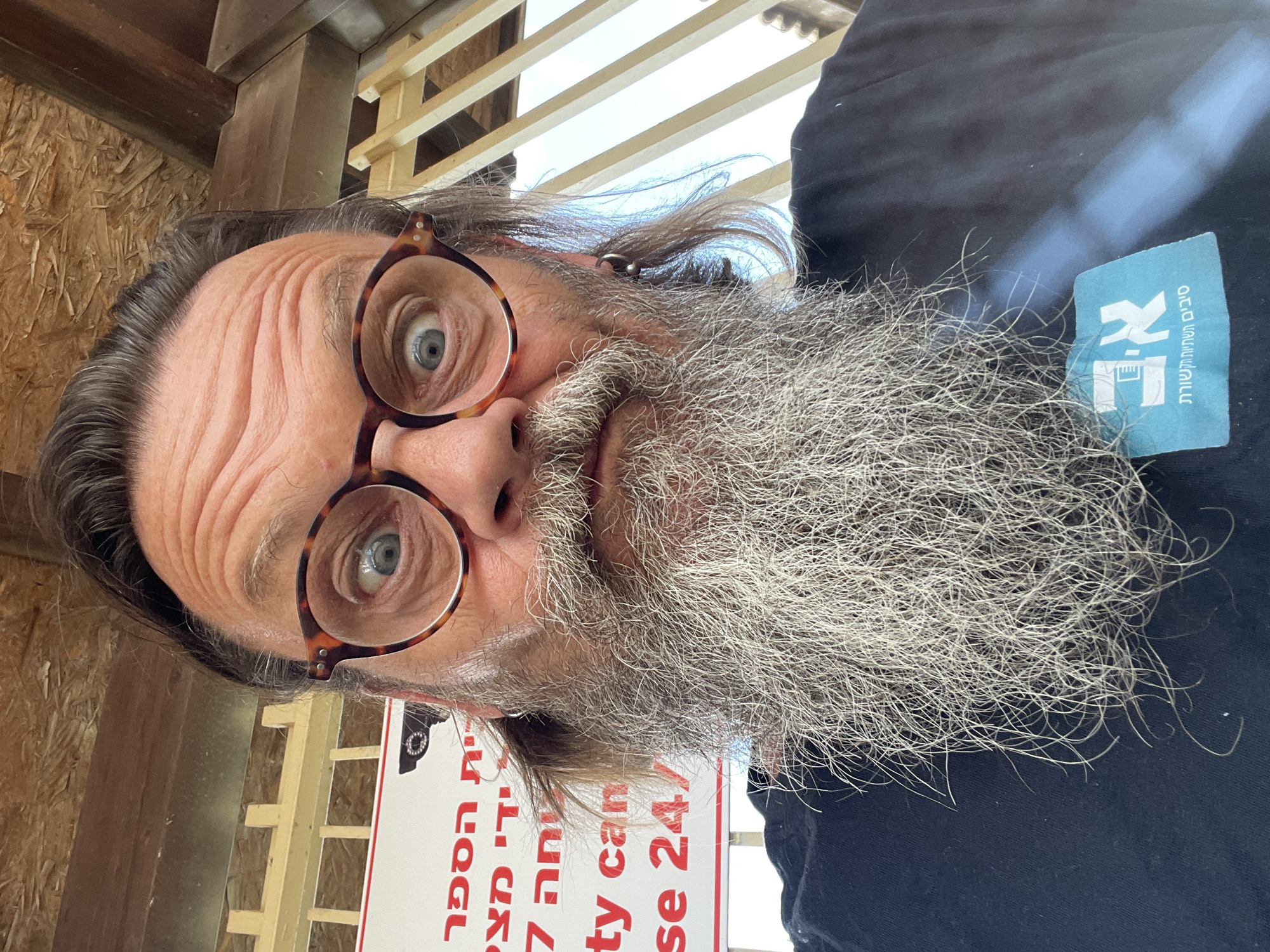 A close-up, selfie-style, eye-level, full-face portrait of a middle-aged, Caucasian male with an extremely long, gray, bushy beard and shoulder-length, dark brown hair. The subject is wearing round, tortoiseshell glasses and a black t-shirt. He has a surprised expression with wide, blue eyes and furrowed brows. A circular silver earring is visible in his left ear. Behind him, a white sign with red lettering and a security camera icon is partially visible. Below the sign, on his t-shirt, is a light blue rectangular patch with white text and a logo. The background consists of light brown wooden panels on the ceiling and a light-colored metal fence. The overall lighting suggests an outdoor environment, possibly in a covered area. The sign appears to be in Hebrew. The location is in the city of Even Yehuda, Israel.
