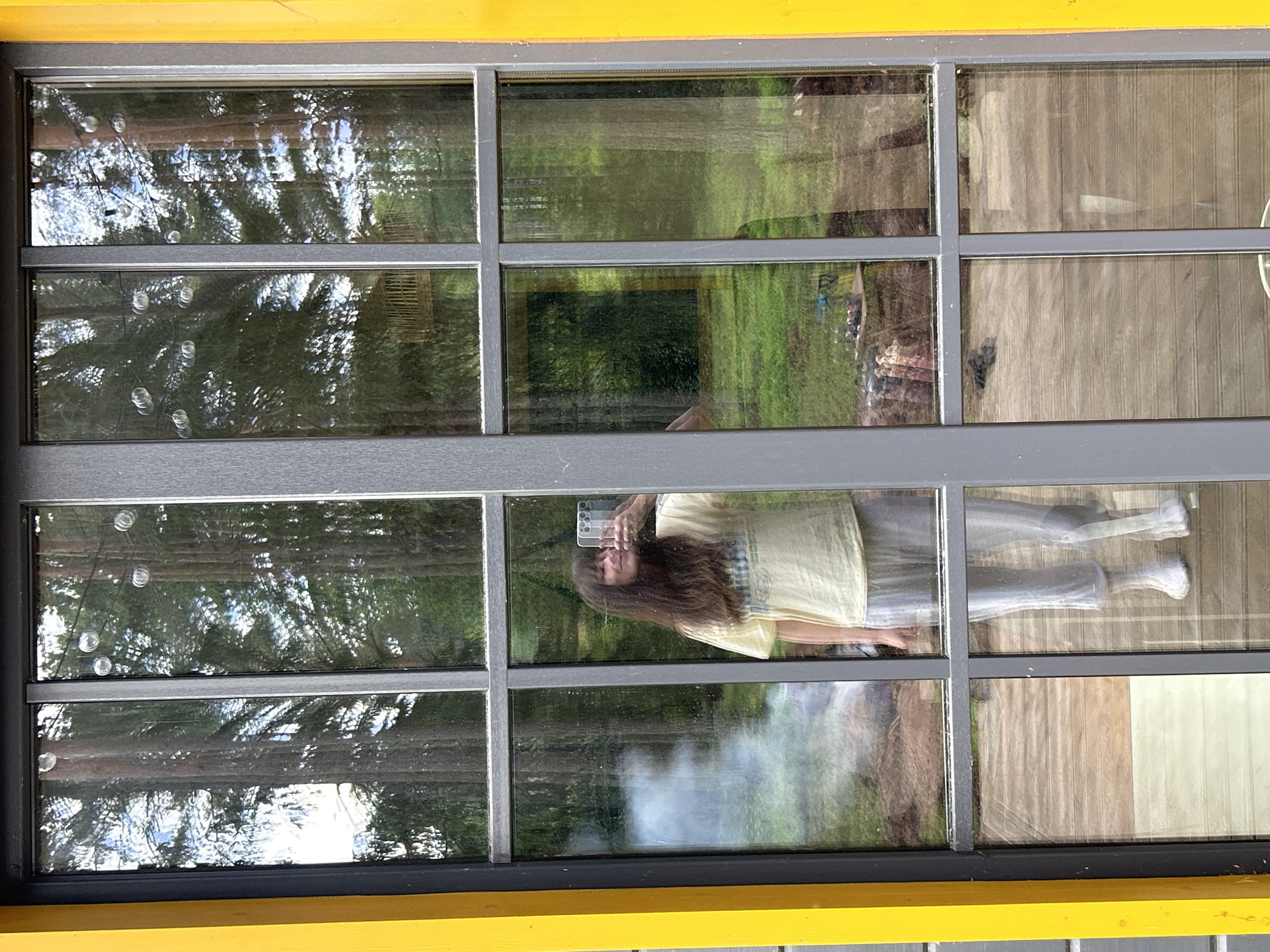 A full-body, mirrored selfie is taken by a person standing indoors and looking out through a large, multi-paned window with yellow framing. The person, with long brown hair and wearing a light yellow t-shirt and gray sweatpants, holds a white smartphone to take the picture. Behind the person, the window reflects a forest scene with tall trees. A faint wispy smoke or fog is visible in the reflection to the left of the person. The foreground shows a wooden deck floor.